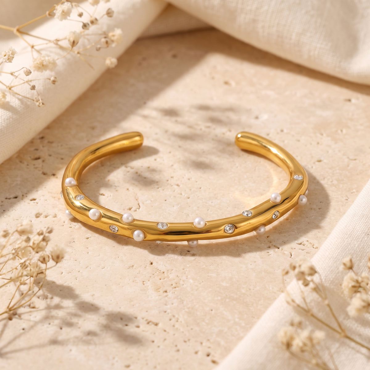 Gold-tone open cuff bracelet with white pearls and cubic zirconia accents on a textured stone surface.