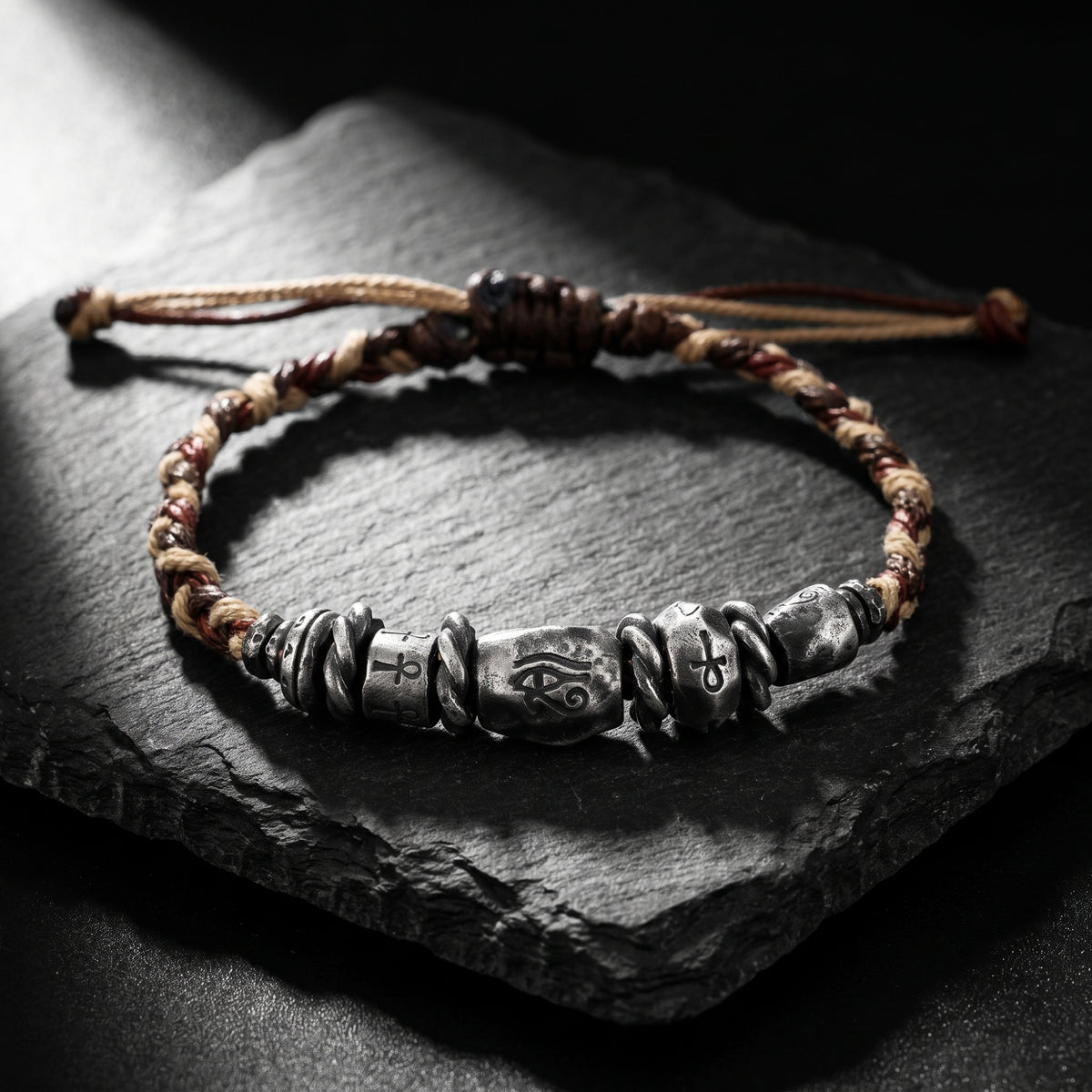 Braided cord bracelet with fine silver Eye of Horus and Ankh beads resting on black slate.