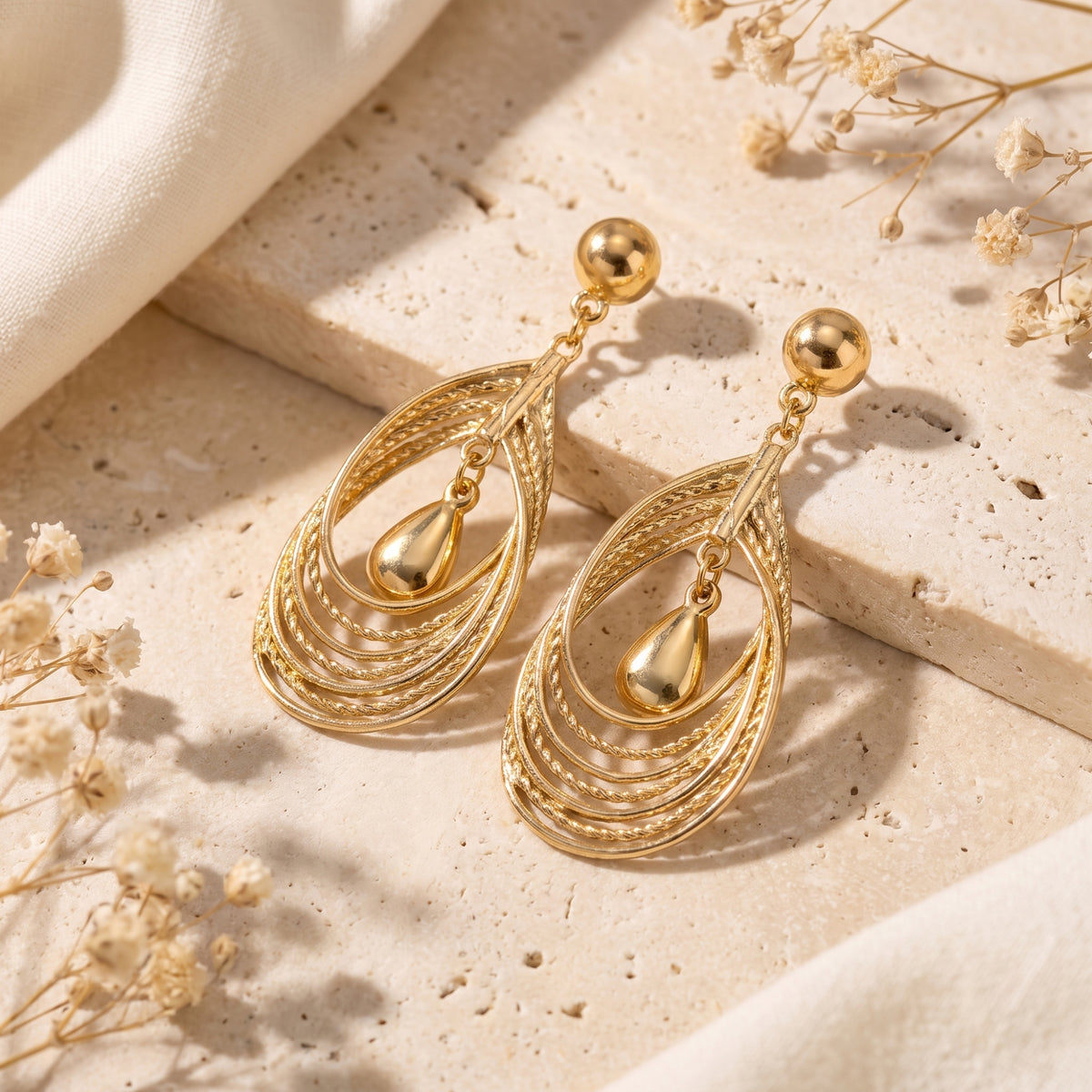 Gold-toned layered teardrop earrings displayed on a beige stone surface with cream fabric and dried flowers.