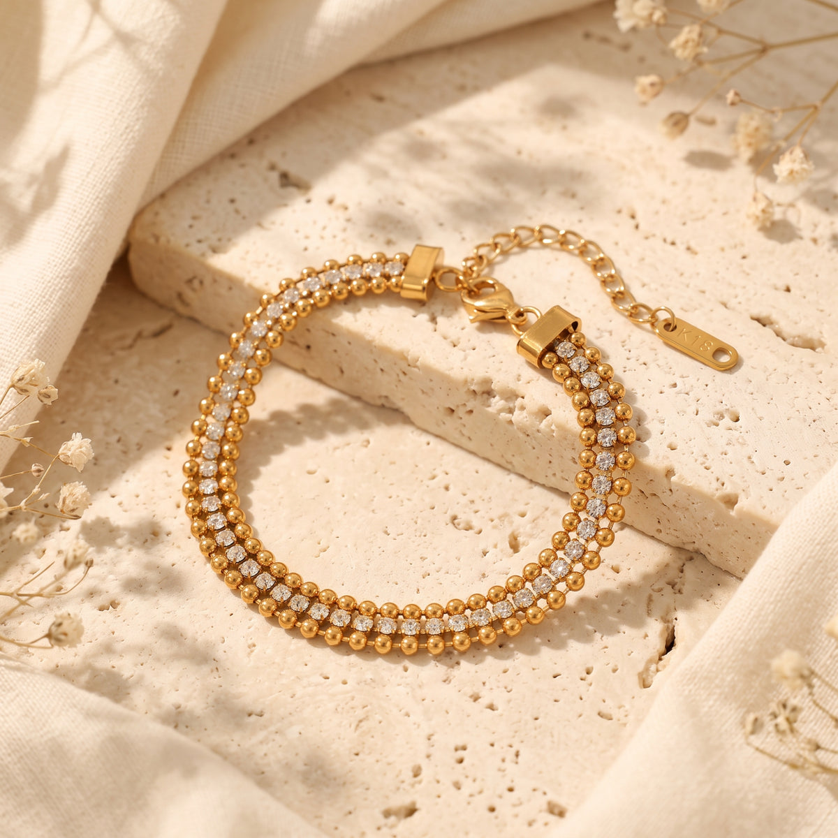 Top-down view of a gold-tone triple-row beaded bracelet with cubic zirconia on a beige stone slab.