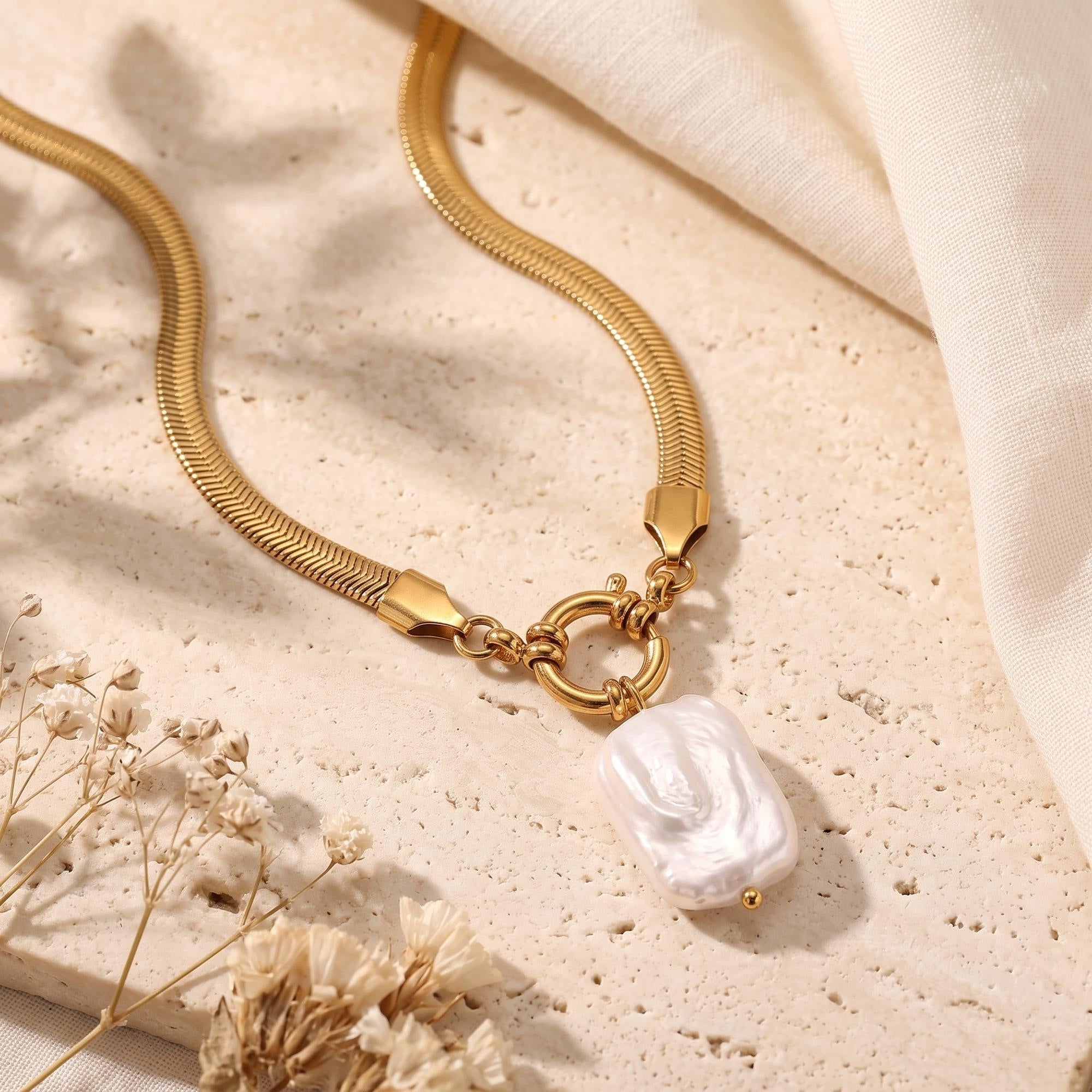 Detail of the gold herringbone necklace clasp and white baroque pearl pendant on a textured stone surface.