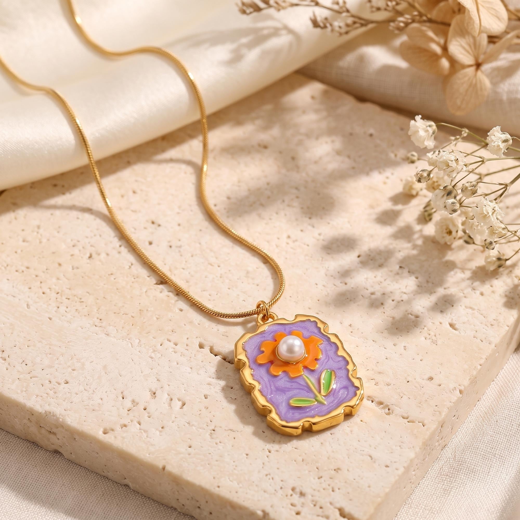 Gold floral necklace with purple enamel and a pearl center styled on a stone surface with dried flowers.