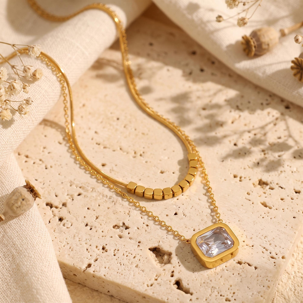 Gold-tone double-layered necklace displayed on a stone slab, with cube-beaded chain and square clear pendant.