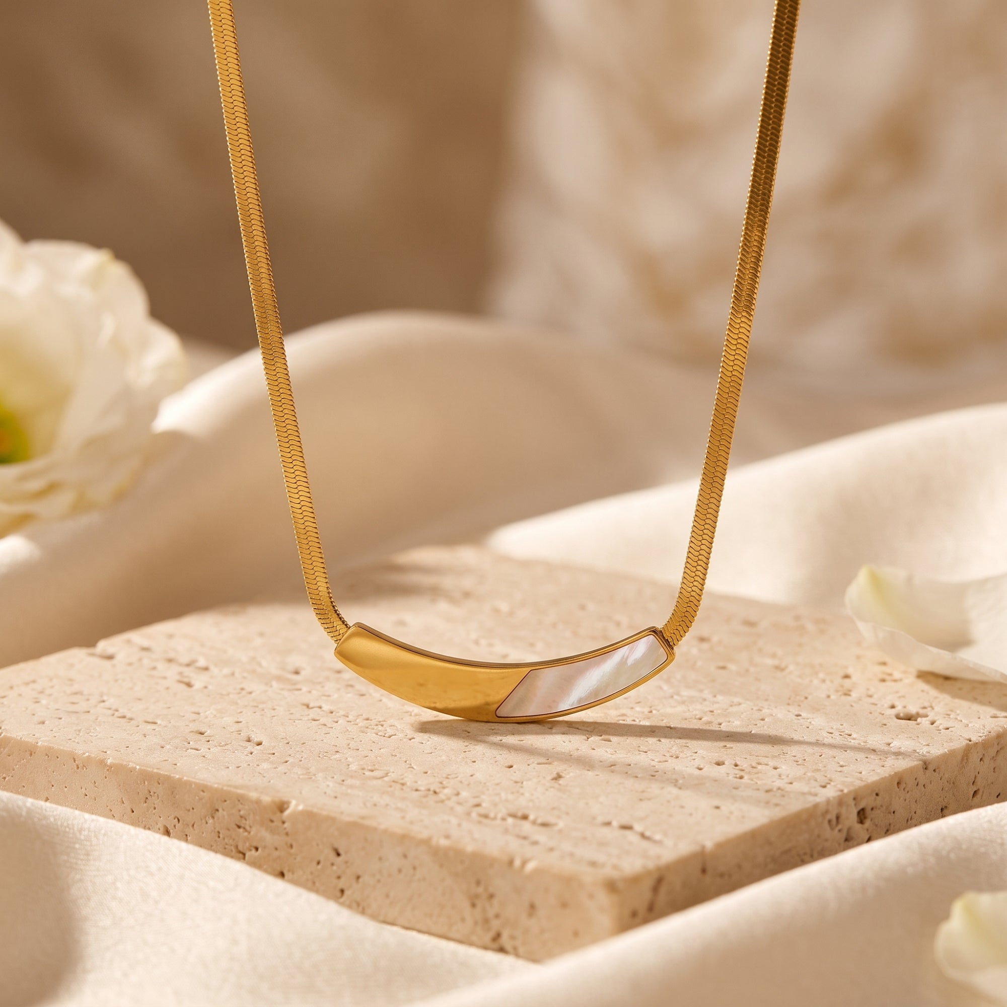 Gold snake chain necklace with mother of pearl inlay on a stone pedestal with white flowers.