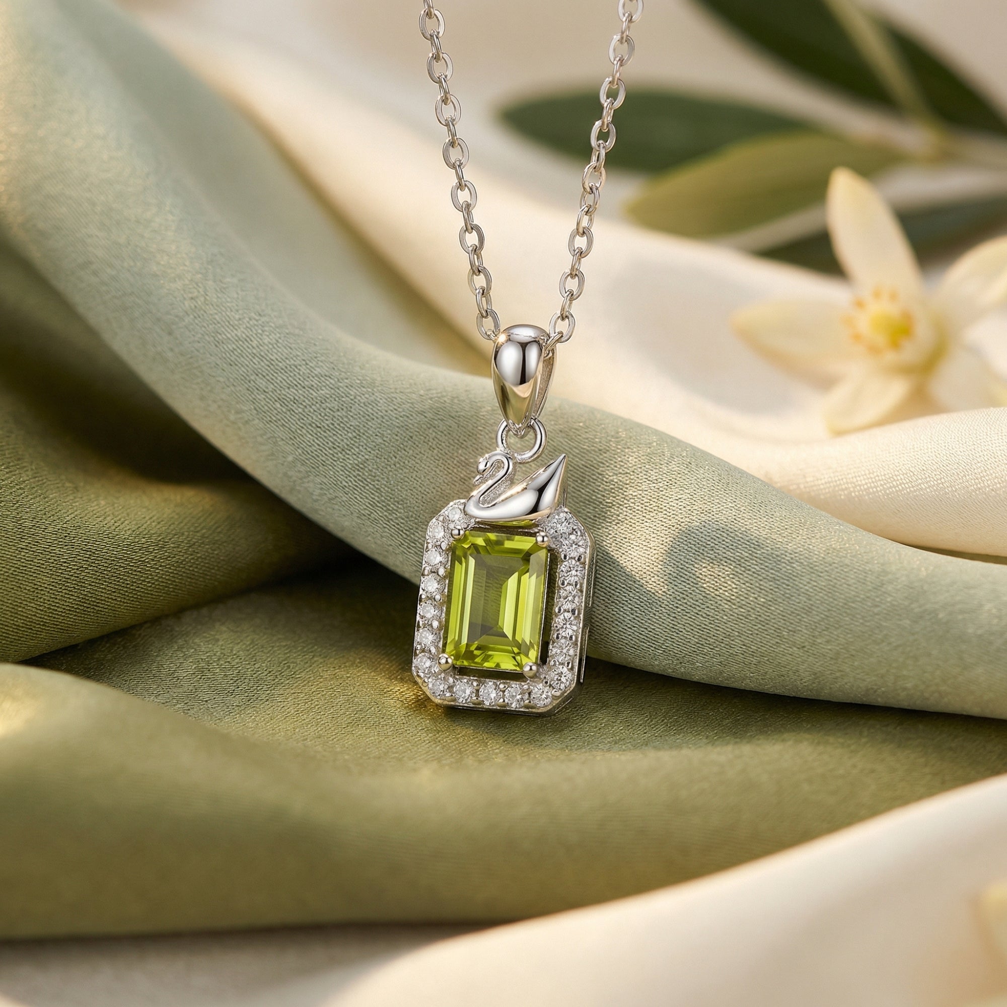 Emerald-cut peridot pendant in S925 sterling silver with a swan motif, resting on draped green and cream silk fabric.