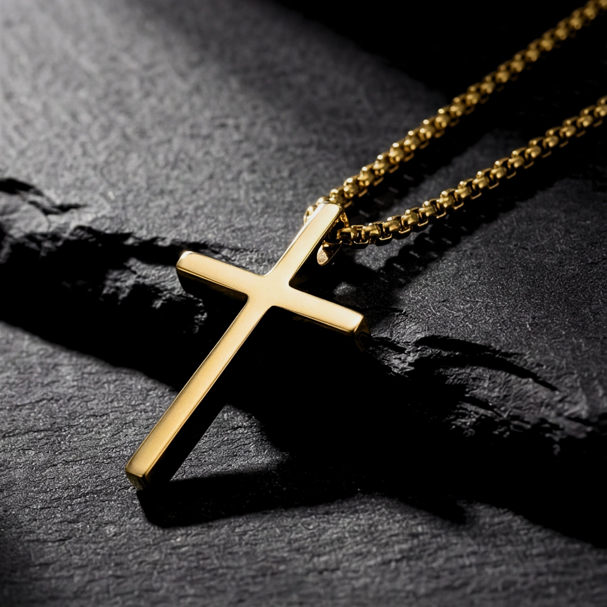 Gold tone stainless steel cross pendant on a box chain, angled view on a dark textured stone surface.
