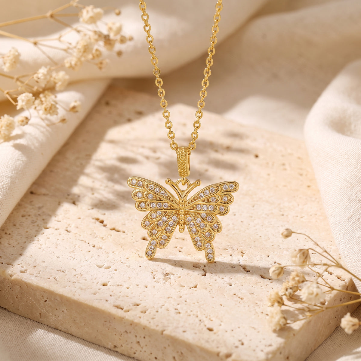 Gold-tone butterfly pendant with clear stones hanging above a beige stone slab with dried white flowers.