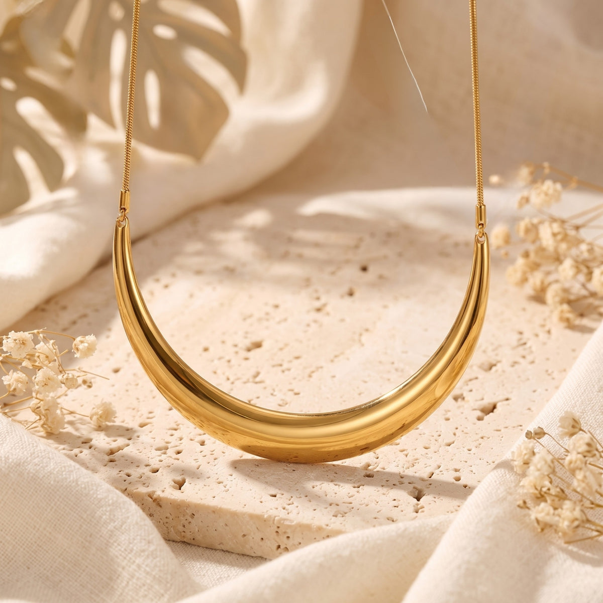 Gold-tone crescent collar necklace displayed on a beige stone surface with white dried flowers.
