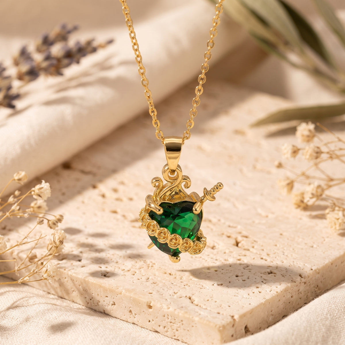 The gold-tone necklace features an emerald green heart pierced by a dagger, suspended over a stone slab.
