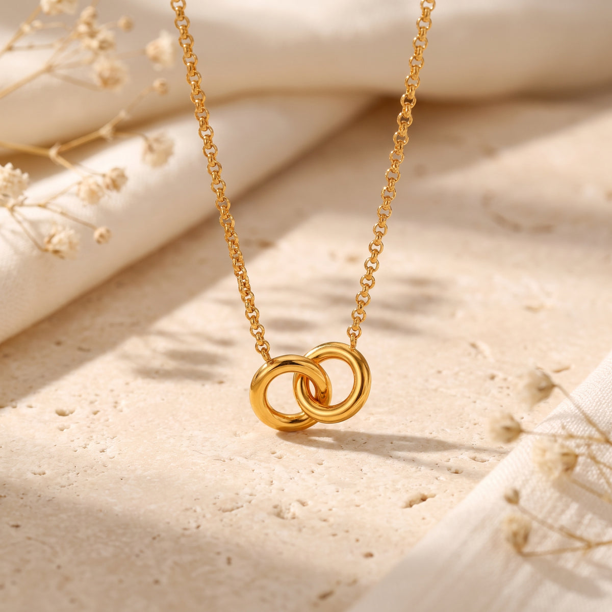Close-up of the gold-tone interlocking ring necklace hanging above stone and cream fabric with dried flowers.