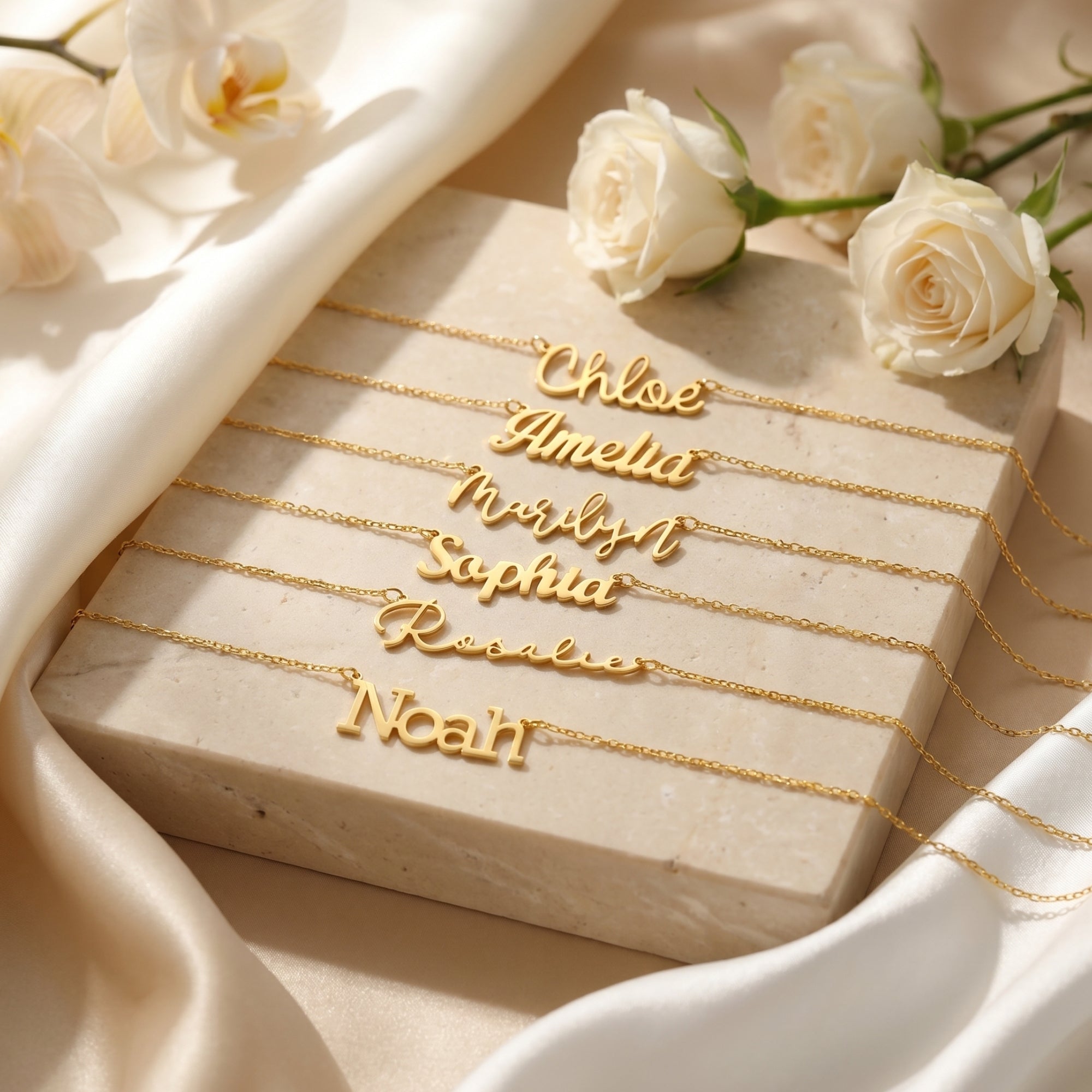 Six 18K gold finish custom name necklaces displayed on a stone block with white roses and silk fabric.
