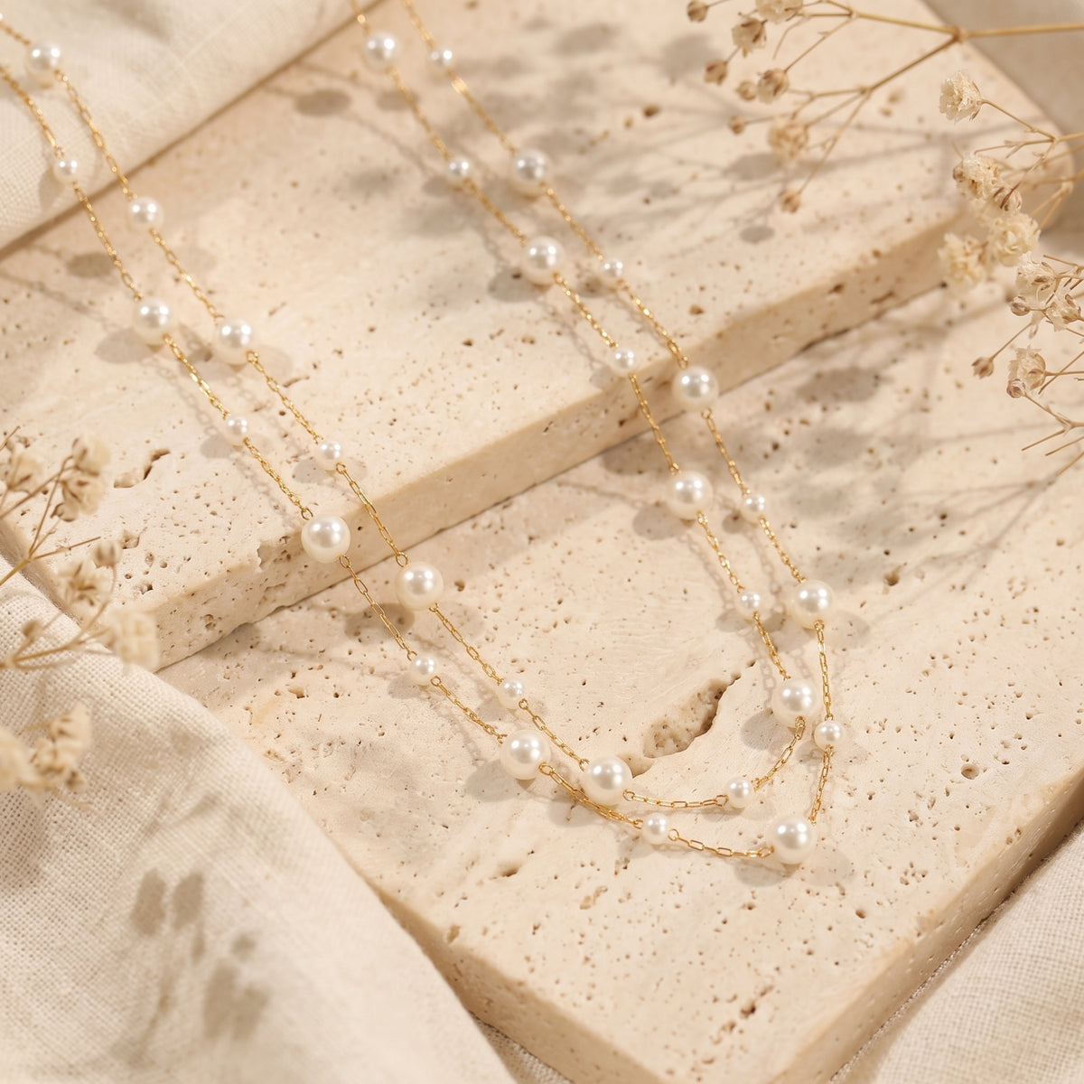 Double-strand gold-tone pearl station necklace displayed on a textured stone surface with dried florals.