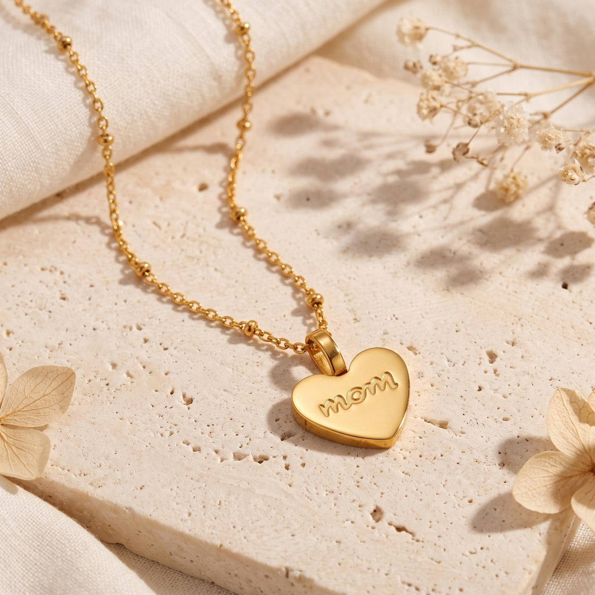 Gold-tone heart pendant engraved with 'mom' on a ball-station chain, resting on a pale stone surface.