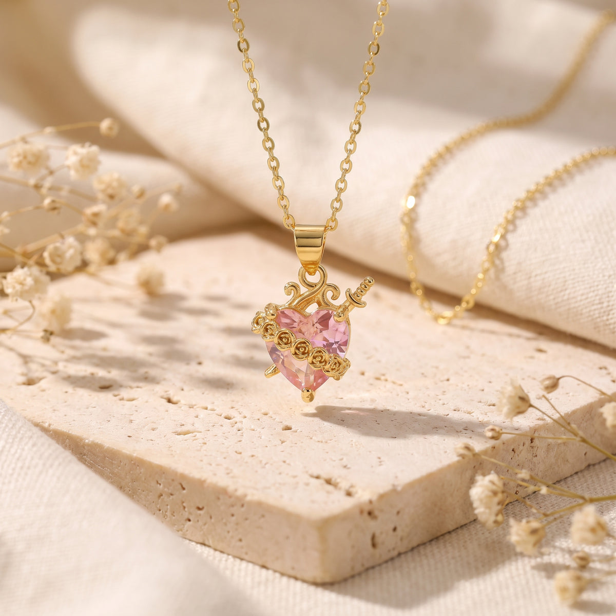 Gold-tone chain hanging vertically with a pink heart pendant pierced by a dagger on a stone tile.