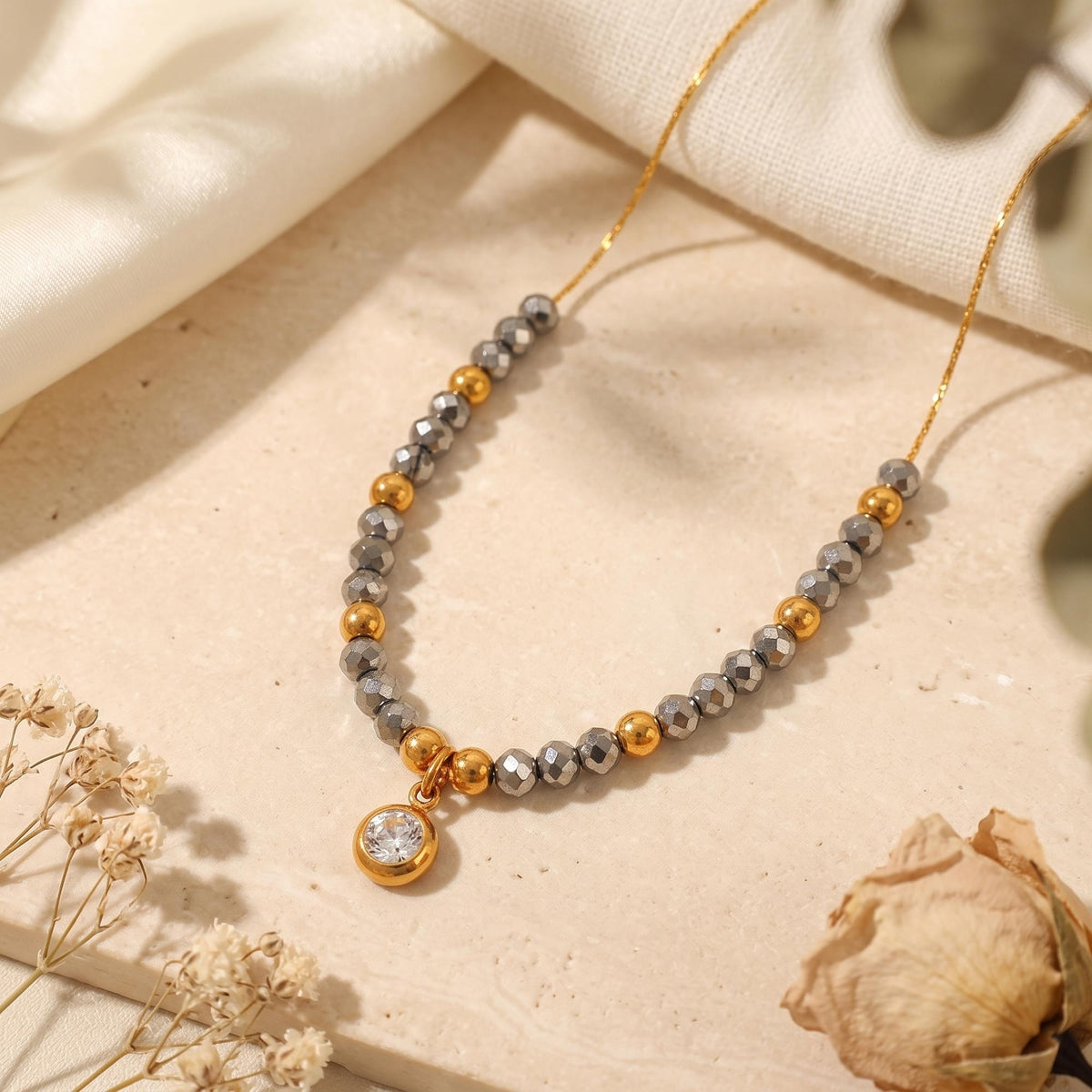Close-up of a gold-tone beaded necklace with faceted gray beads and a round clear pendant on a light stone surface.
