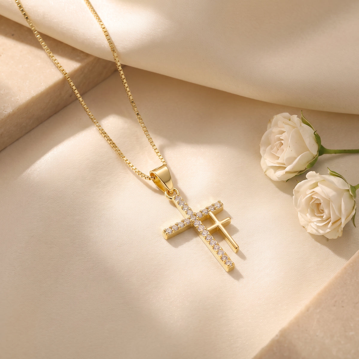 Gold-tone double cross necklace with cubic zirconia accents on cream fabric beside white roses