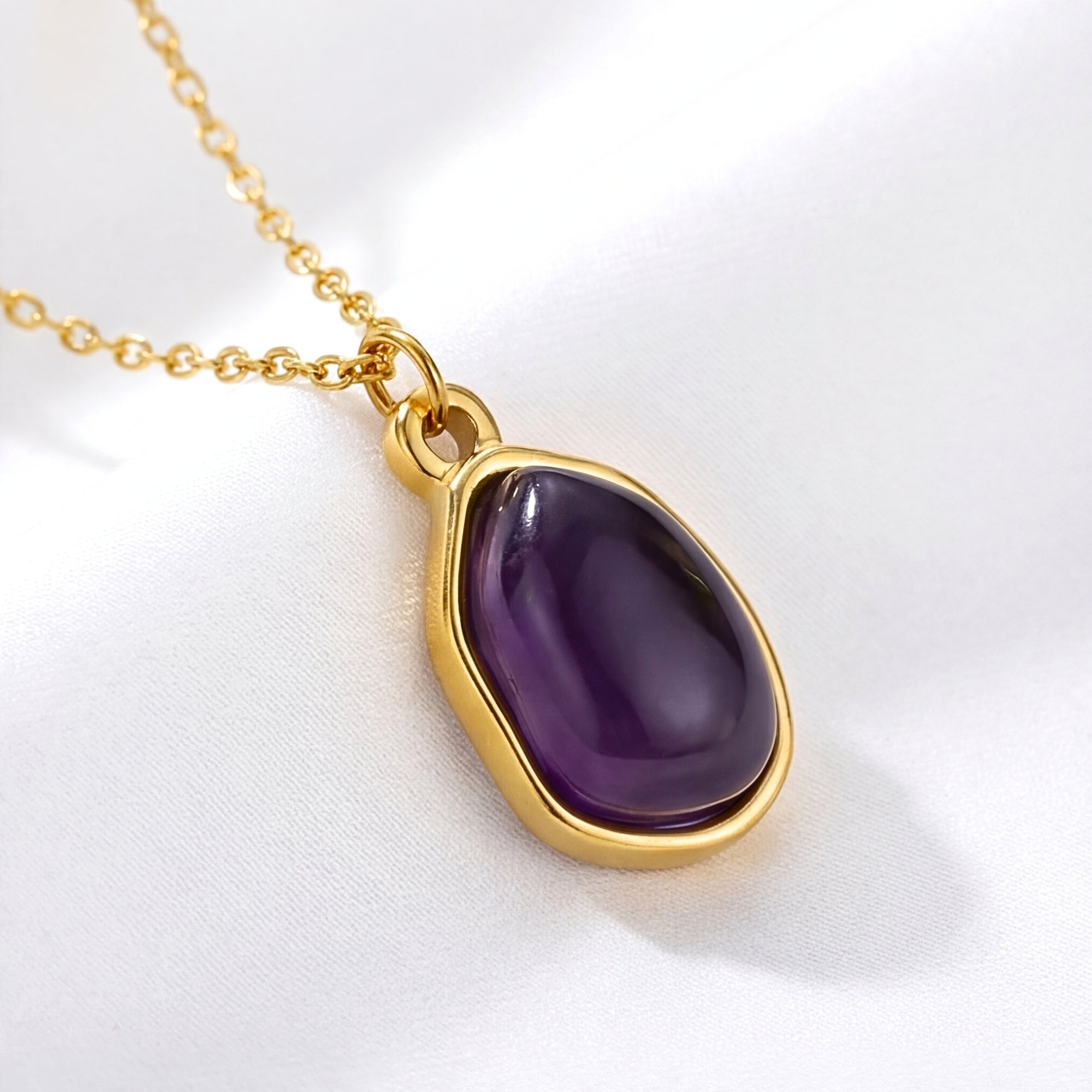 Teardrop simulated amethyst necklace in yellow gold resting on a draped white fabric background.