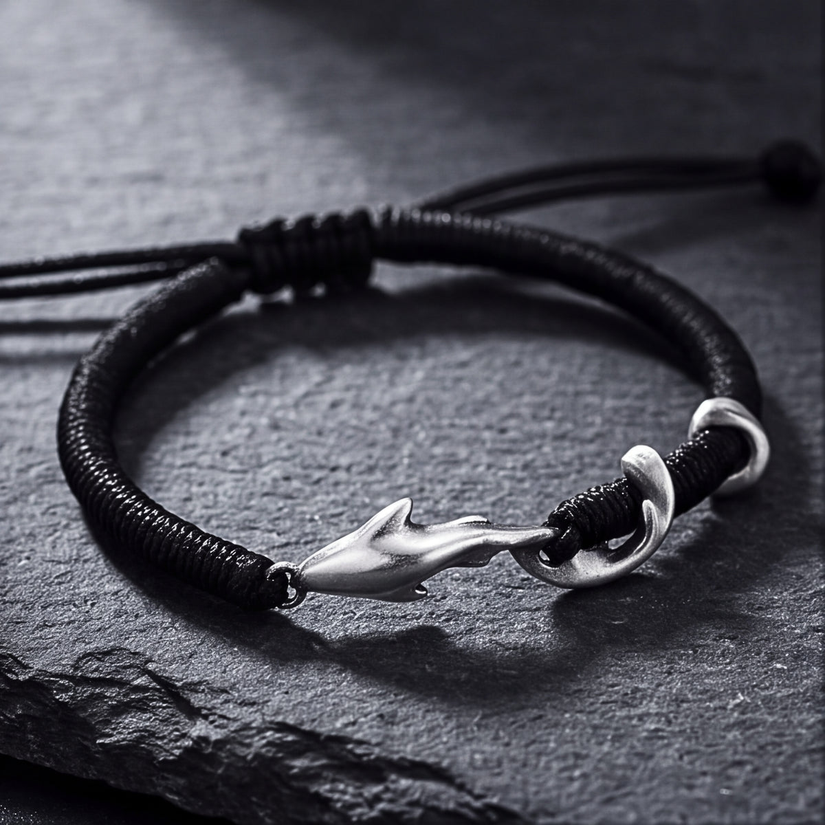 Black cord bracelet with a silver dolphin hook clasp resting flat on a dark slate surface.