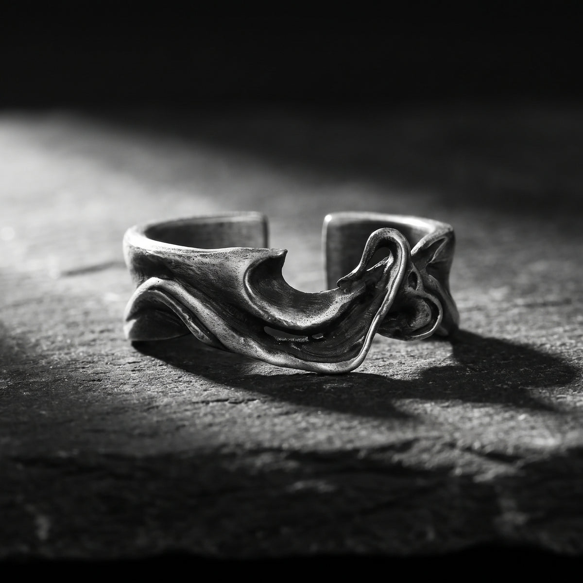 Sculpted silver wave open ring resting on a rough dark stone surface in dramatic light.