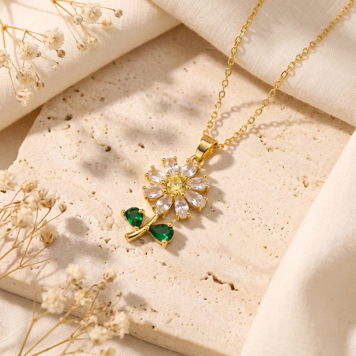 Close-up of a gold-tone daisy pendant necklace with clear, yellow, and green stones resting on a beige stone slab.
