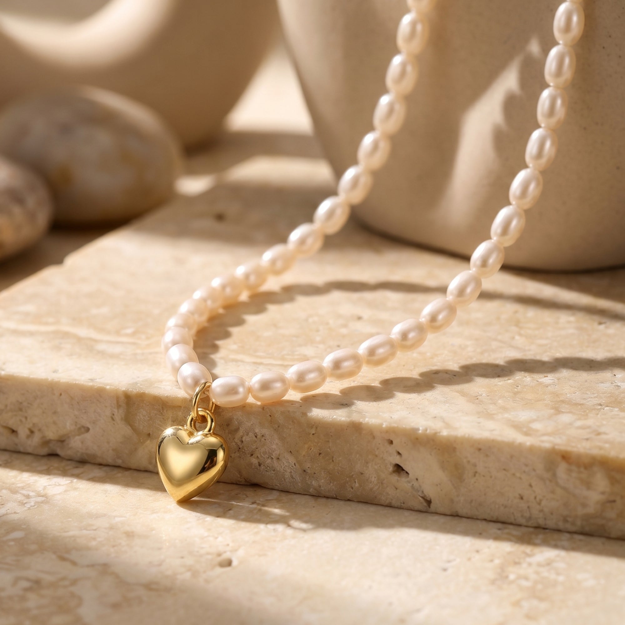 Macro view of The Colette Pearl Necklace, featuring a solid yellow gold heart pendant on a white synthetic pearl necklace on a stone surface.