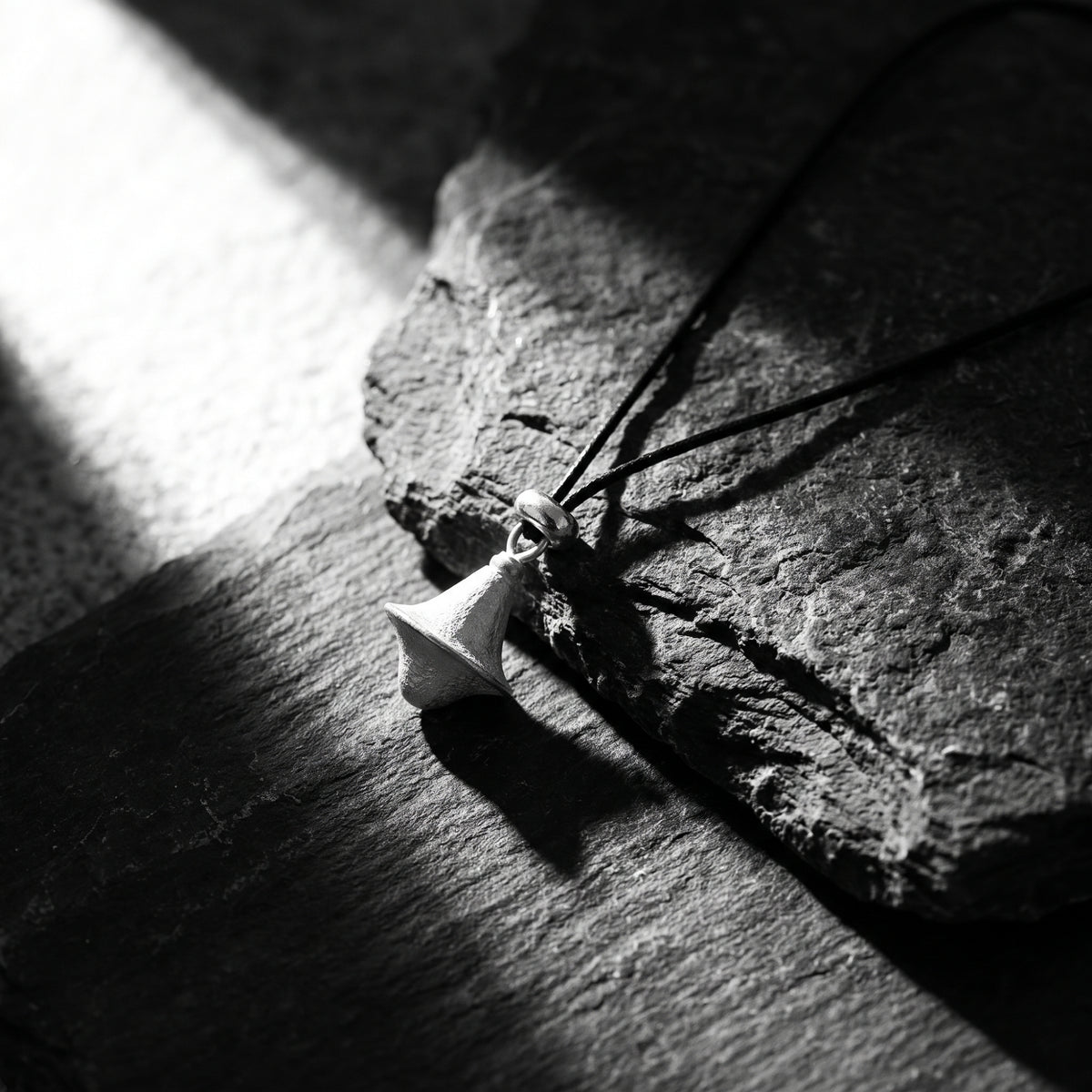 Textured silver double-cone pendant on a black cord resting on dark slate in high-contrast light.