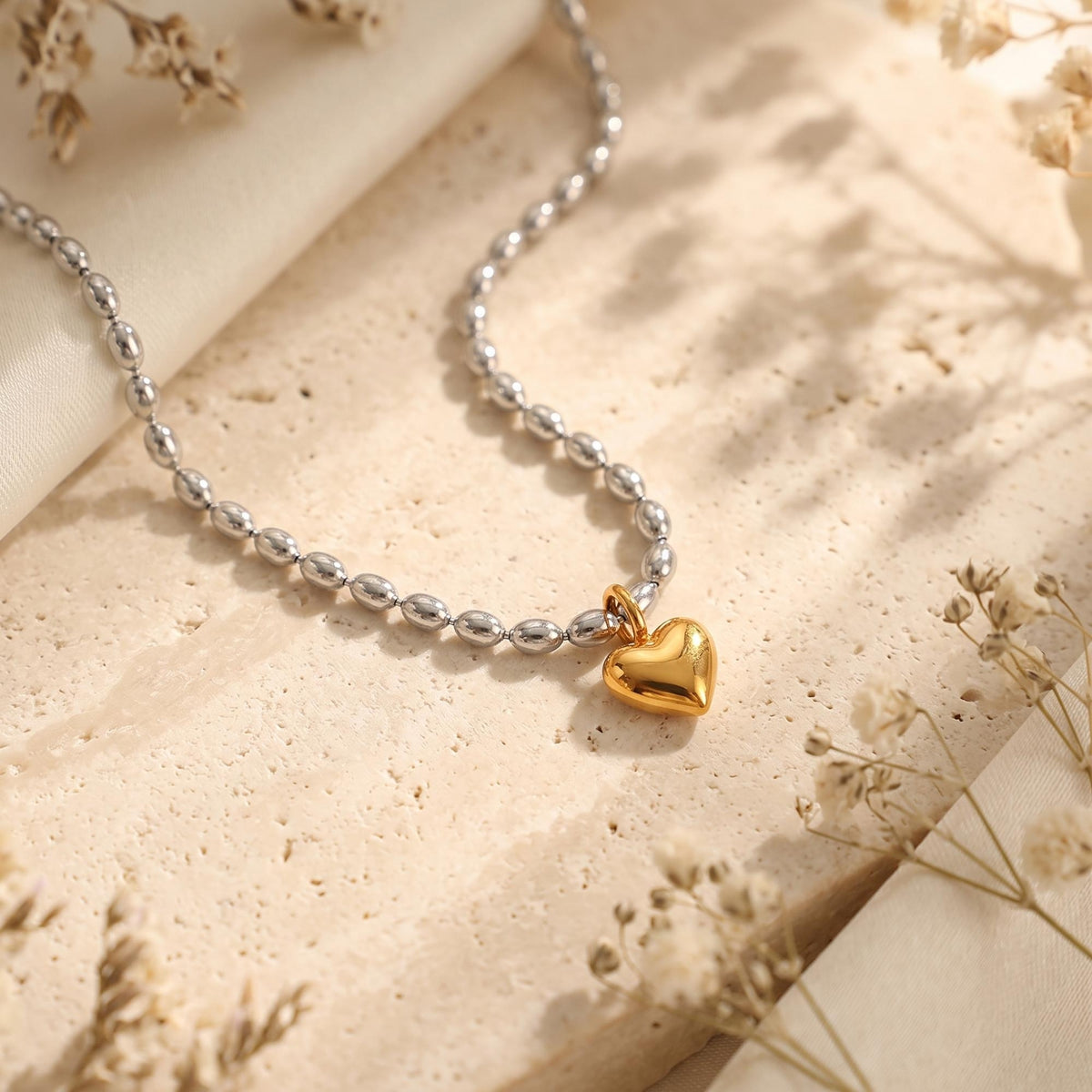Close-up of a gold-tone puffed heart pendant on a silver-tone oval bead chain over a beige stone surface.