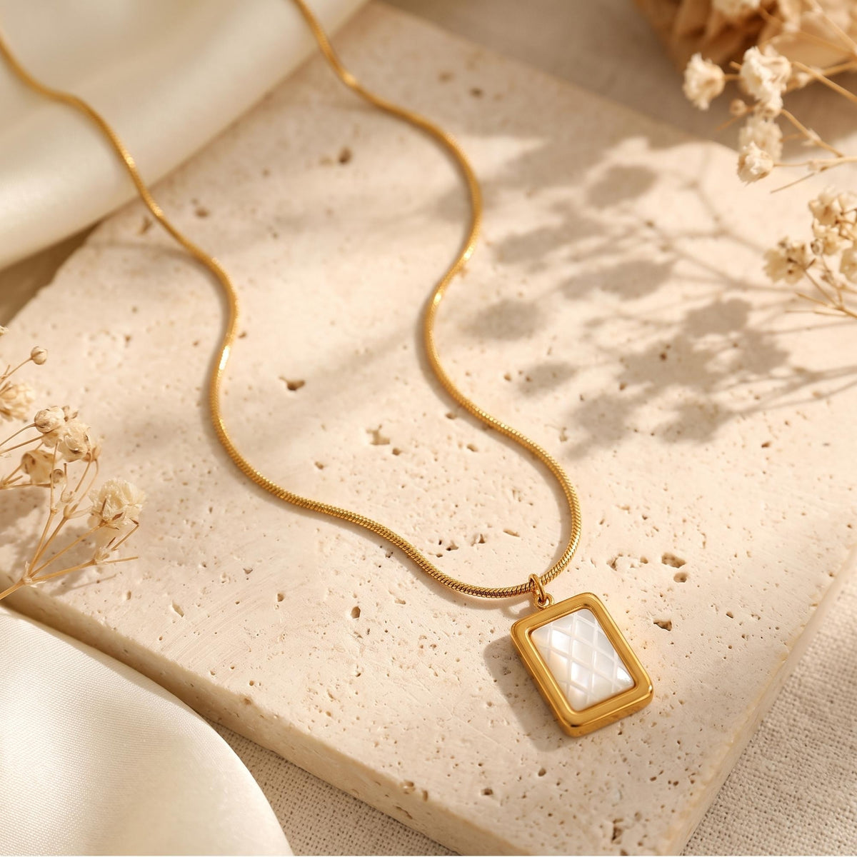 Gold-tone snake chain necklace with a rectangular gold-tone pendant and white quilted inlay on a porous stone slab.