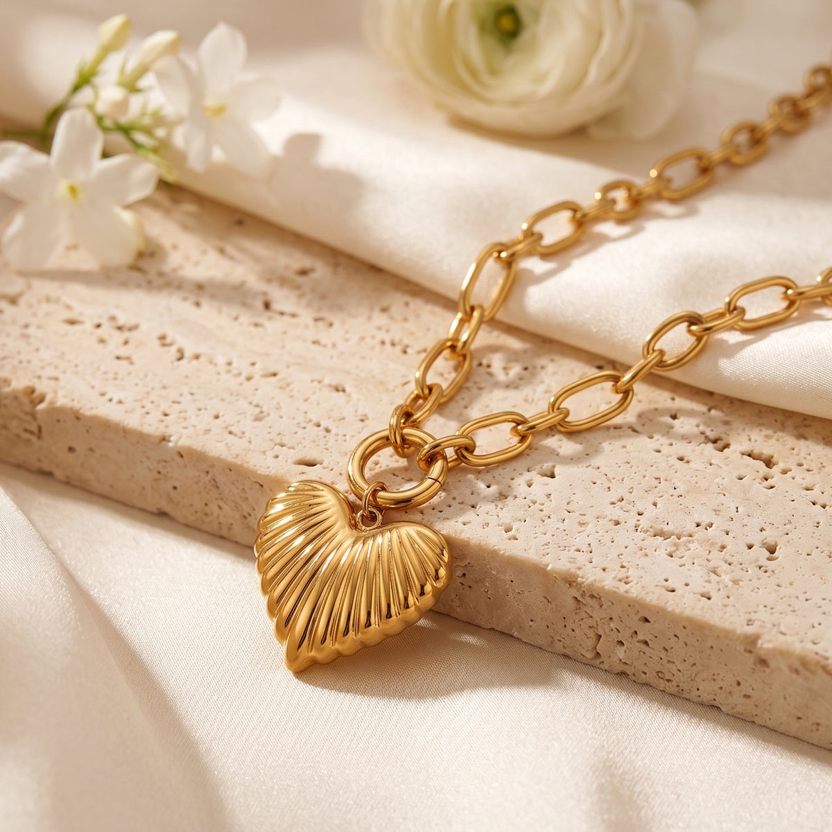 Gold-tone ribbed heart pendant necklace with link chain displayed on a stone slab with white flowers.