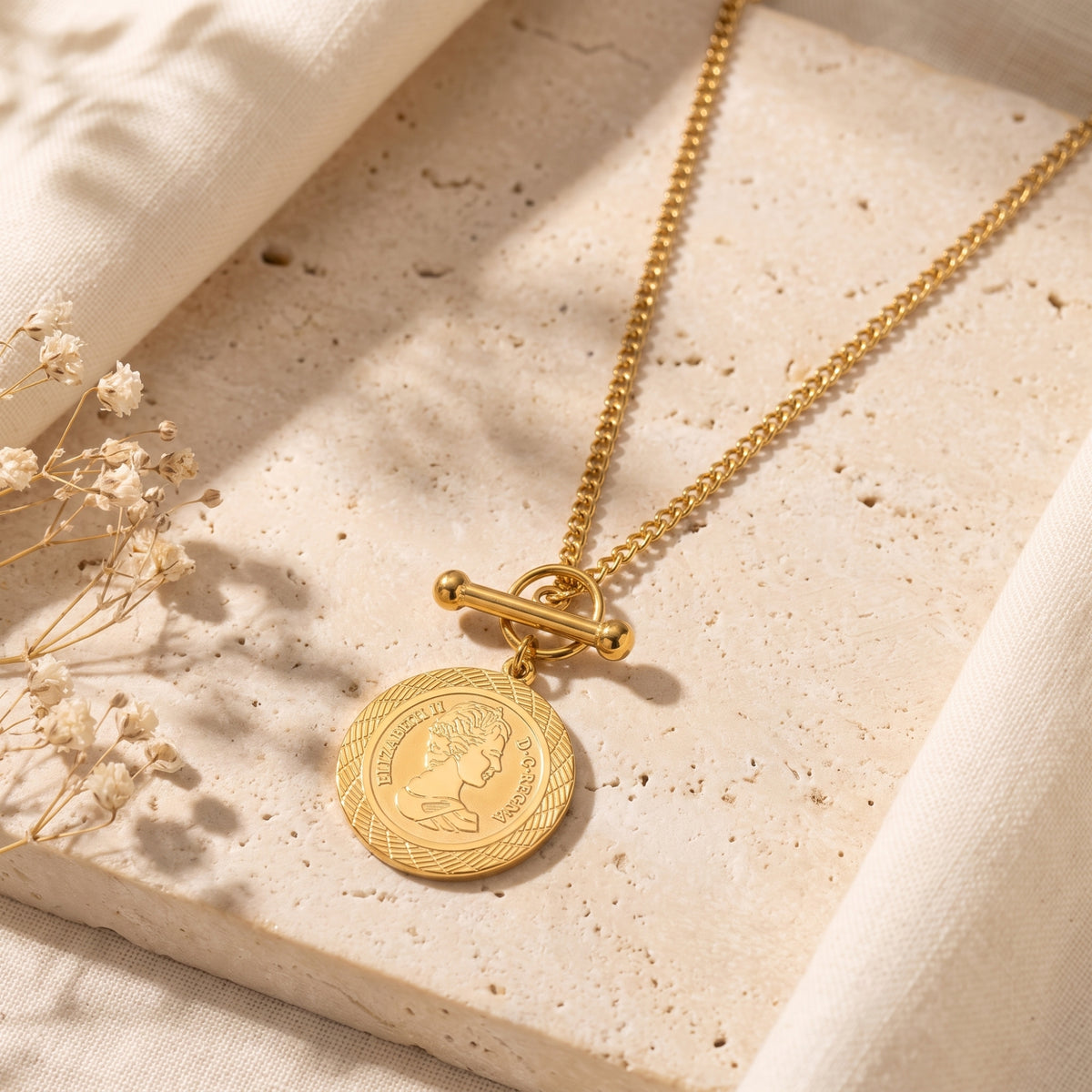 Gold-tone coin medallion necklace with a front T-bar clasp resting on a textured stone surface with dried flowers.