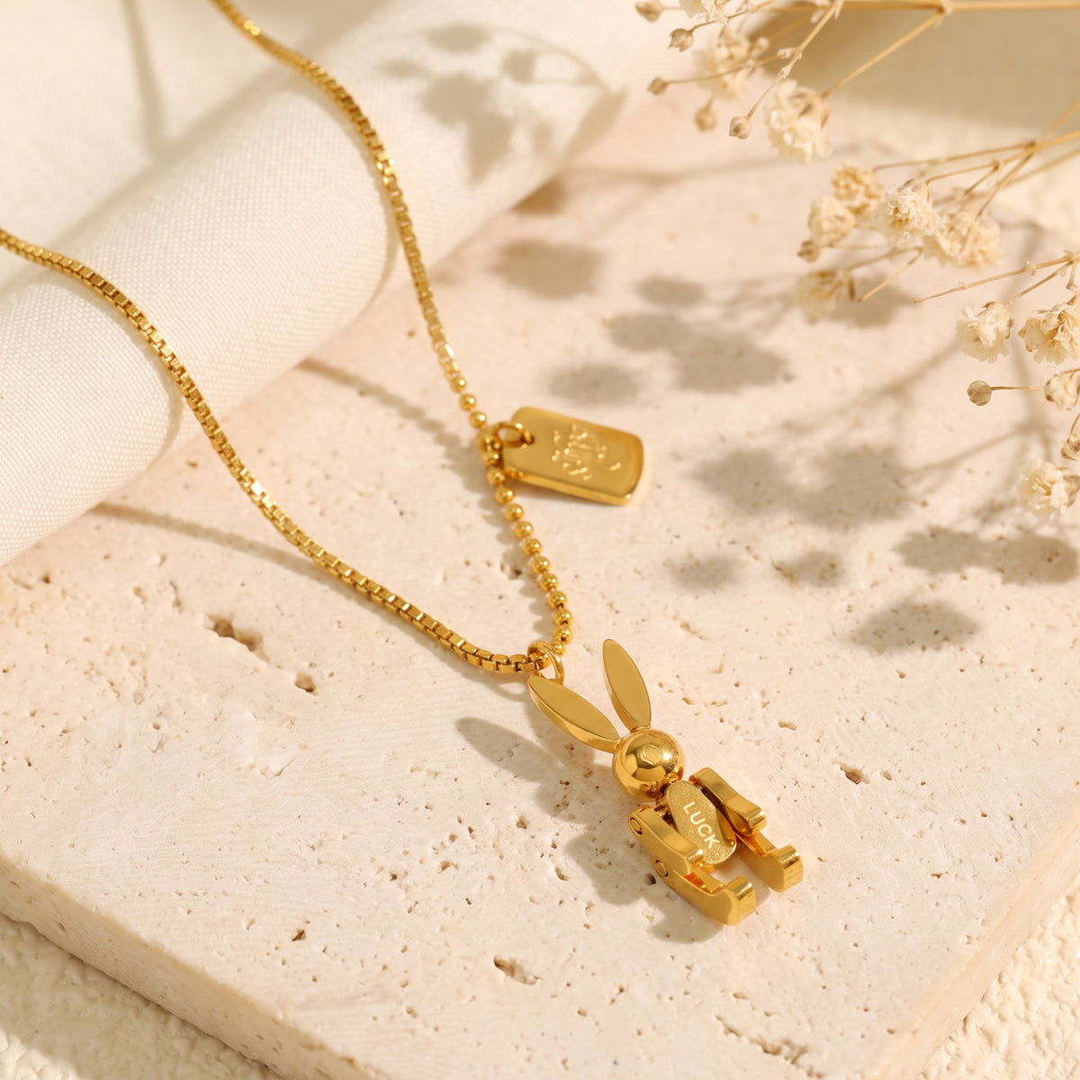 Gold articulated rabbit pendant necklace displayed on a textured beige stone slab with dried flowers.