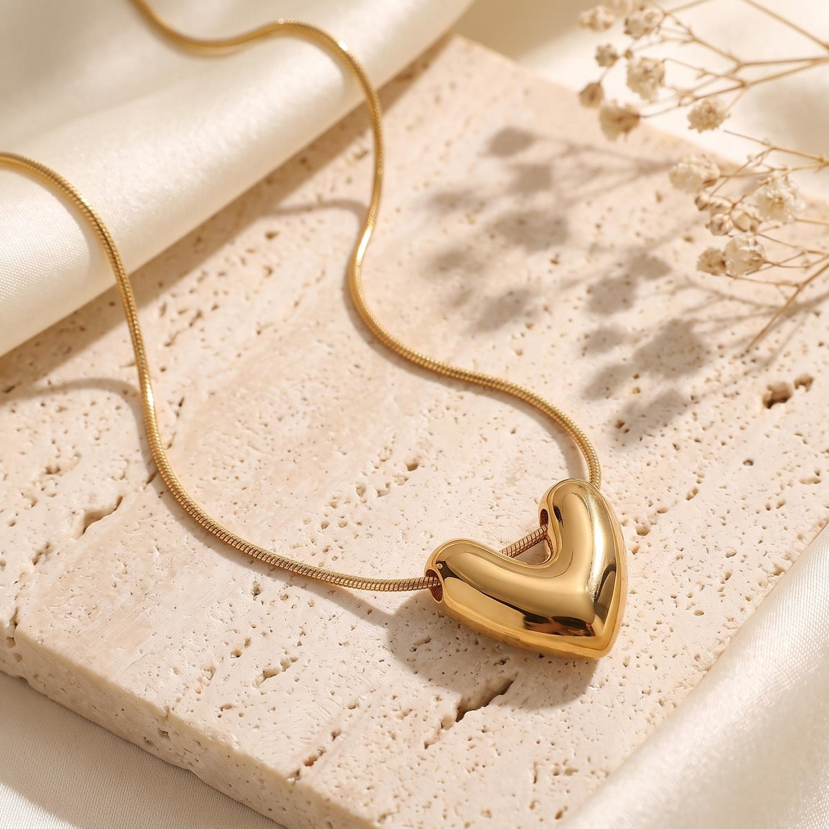 Gold-tone snake chain with a thick sculpted heart pendant on a textured stone surface beside cream fabric.