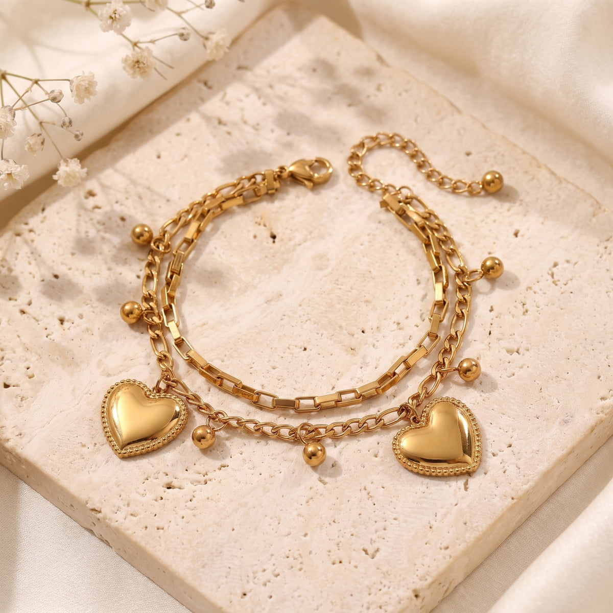 Overhead view of a gold-tone double-chain bracelet with two heart charms and bead drops on a stone tray.