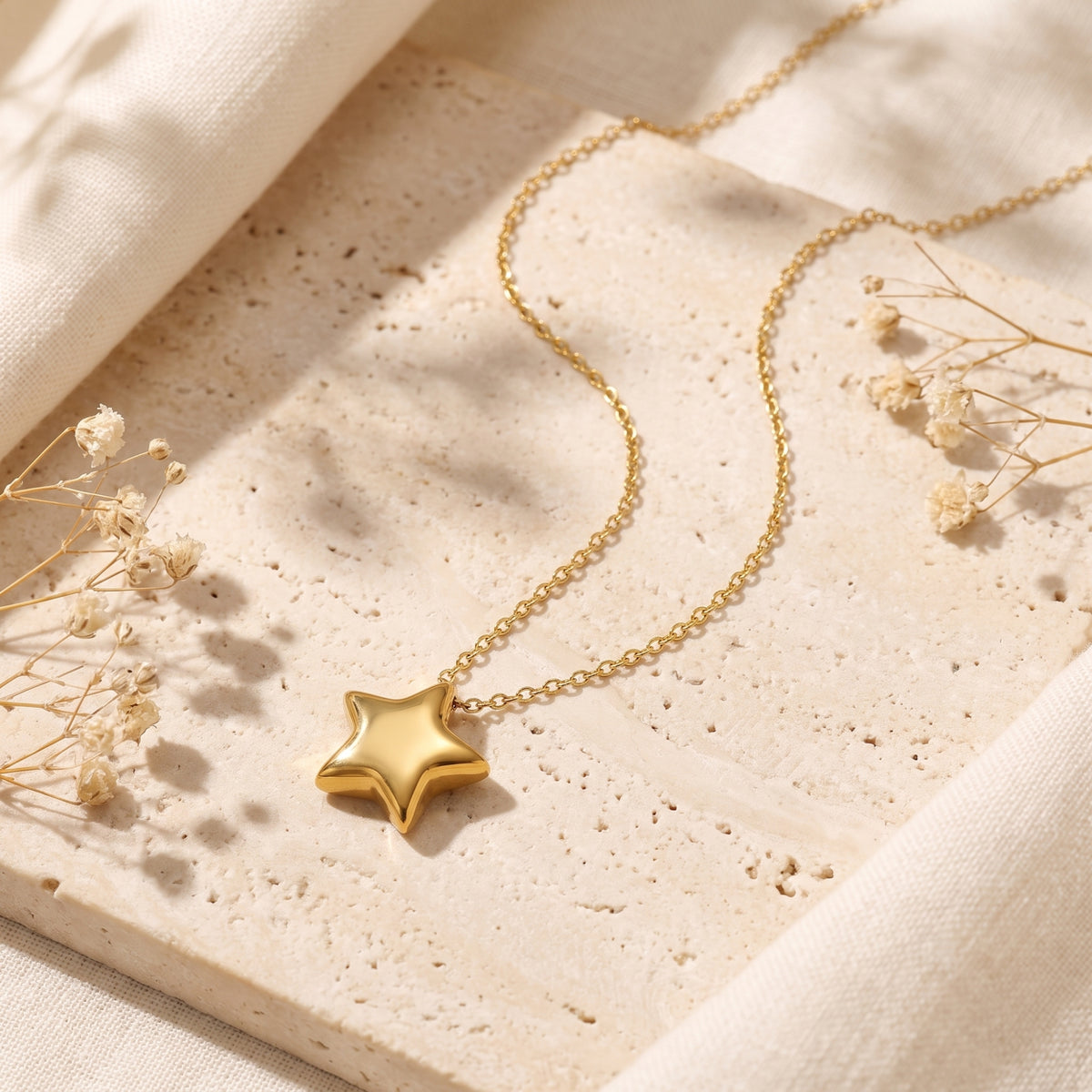Gold-tone 3D puffed star pendant necklace laid flat on a textured stone tile with dried flowers.