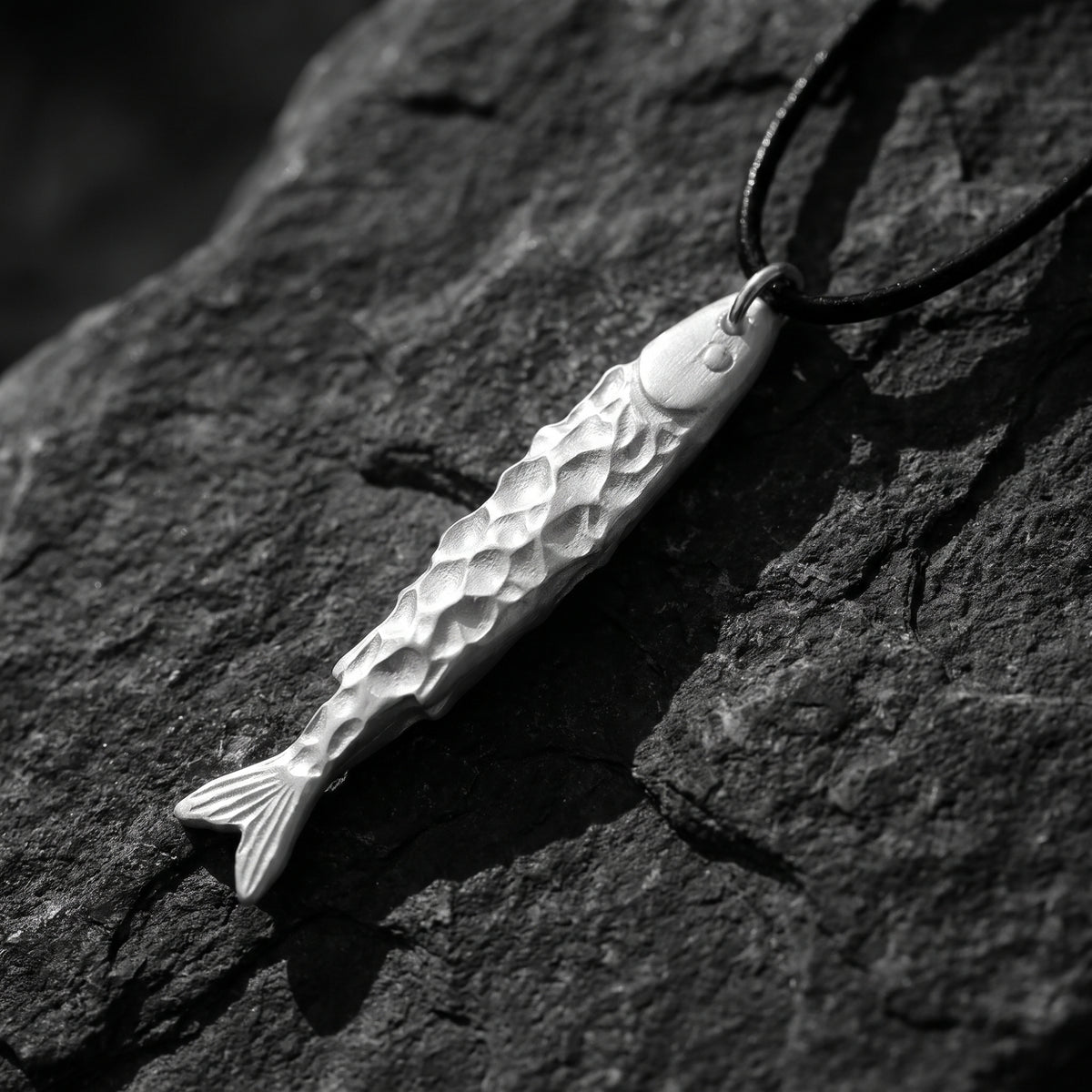 Close-up of a silver textured fish pendant resting on dark slate rock on a black cord.
