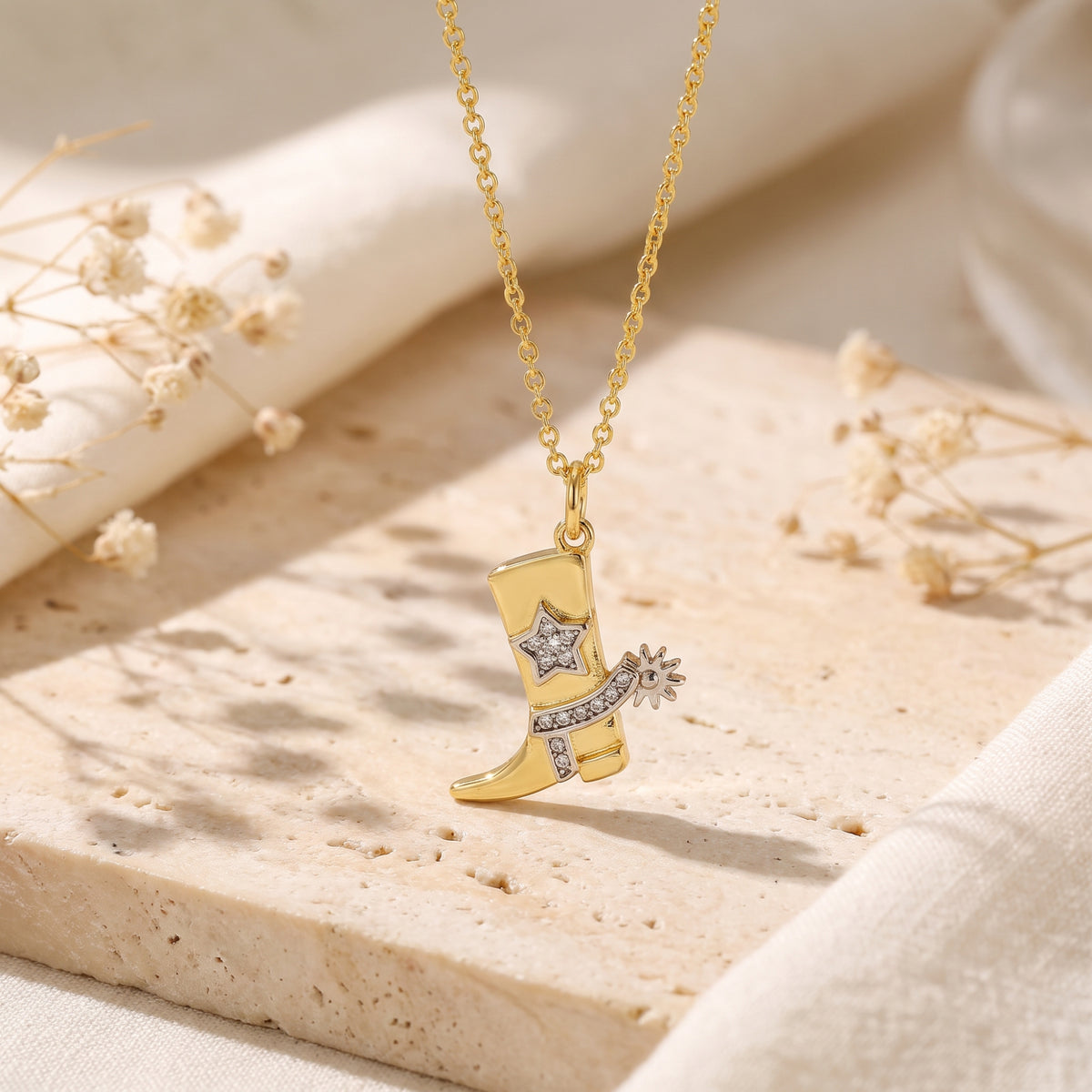 Gold-tone cowboy boot pendant with a clear stone star and spur details, displayed close-up on a textured stone slab.