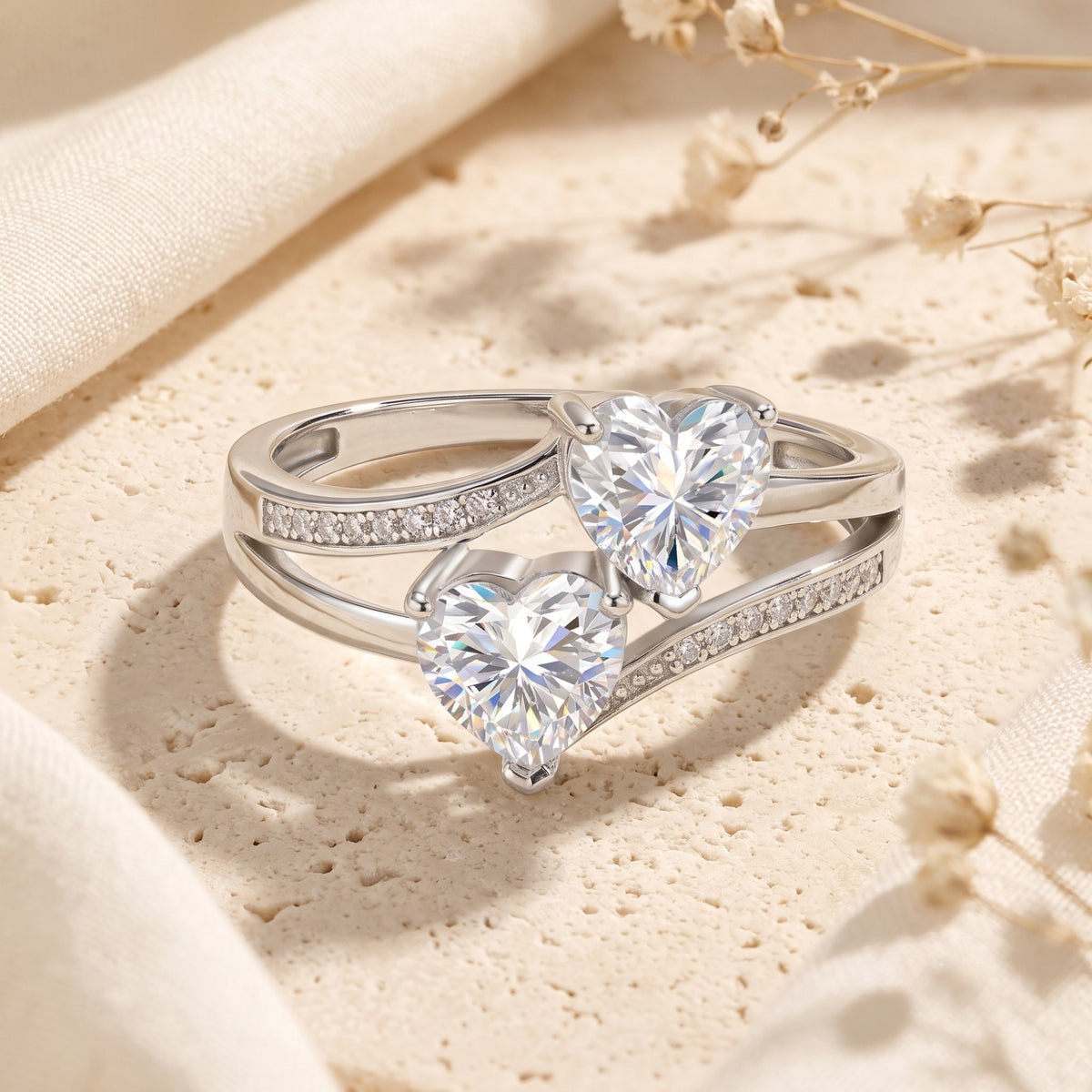 Silver bypass ring with twin heart-cut clear stones and pavé accents displayed flat on textured beige stone.