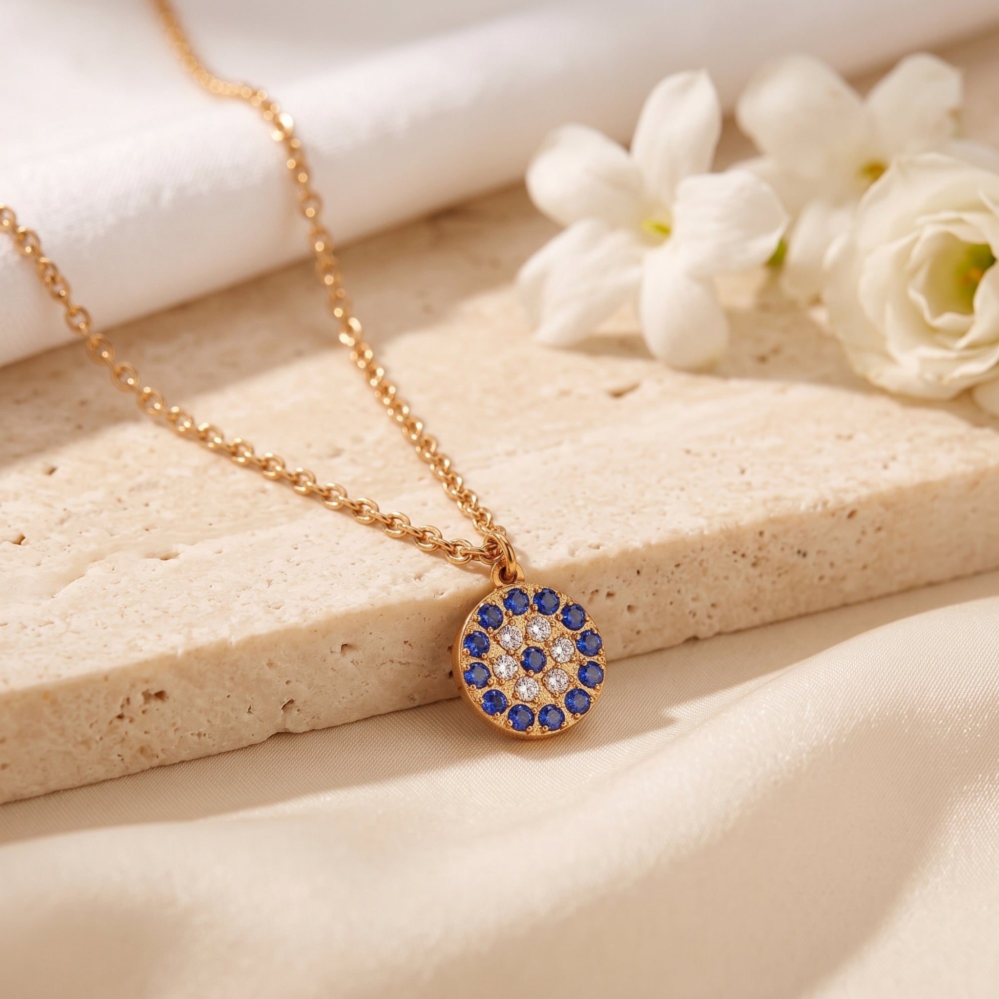 Gold PVD pendant necklace with blue and white pave sapphires on a stone slab with white flowers.