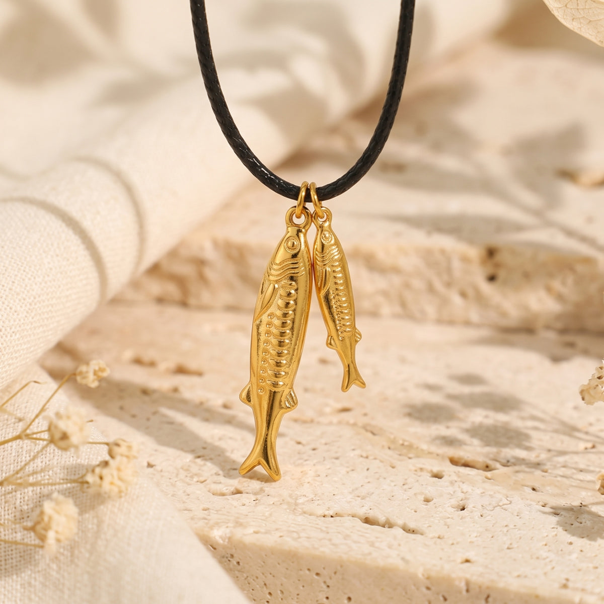 Close-up of two detailed gold-tone fish pendants suspended on a black cord over a textured stone surface.