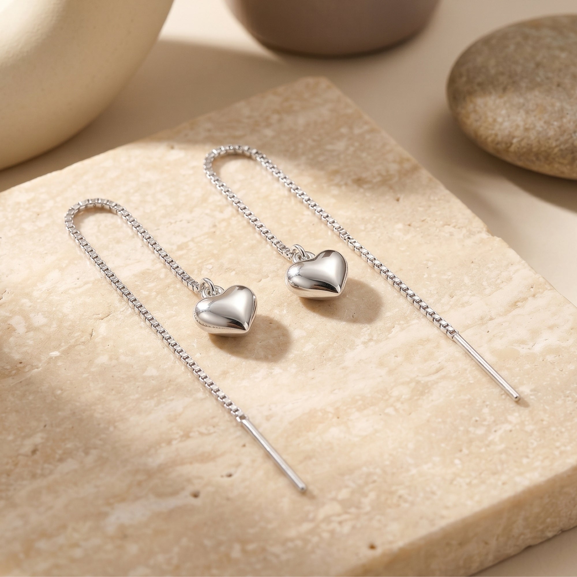 Silver heart threader earrings with box chains displayed on a textured stone surface in a lifestyle setting.