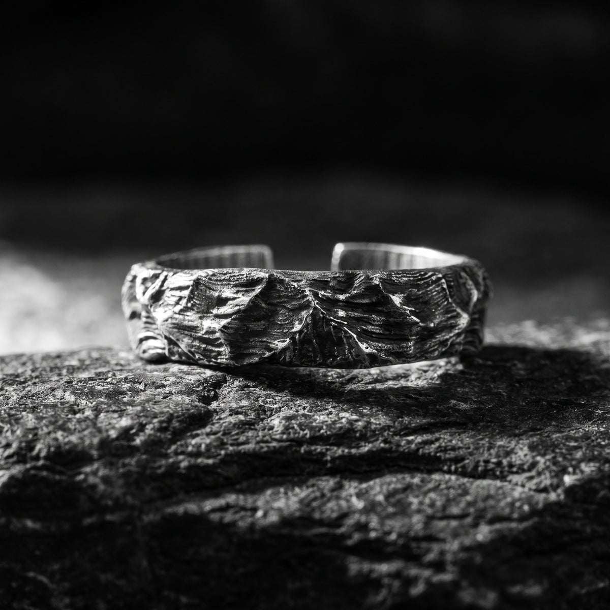 Front view of a silver open-band ring with carved terrain texture resting on dark rock.