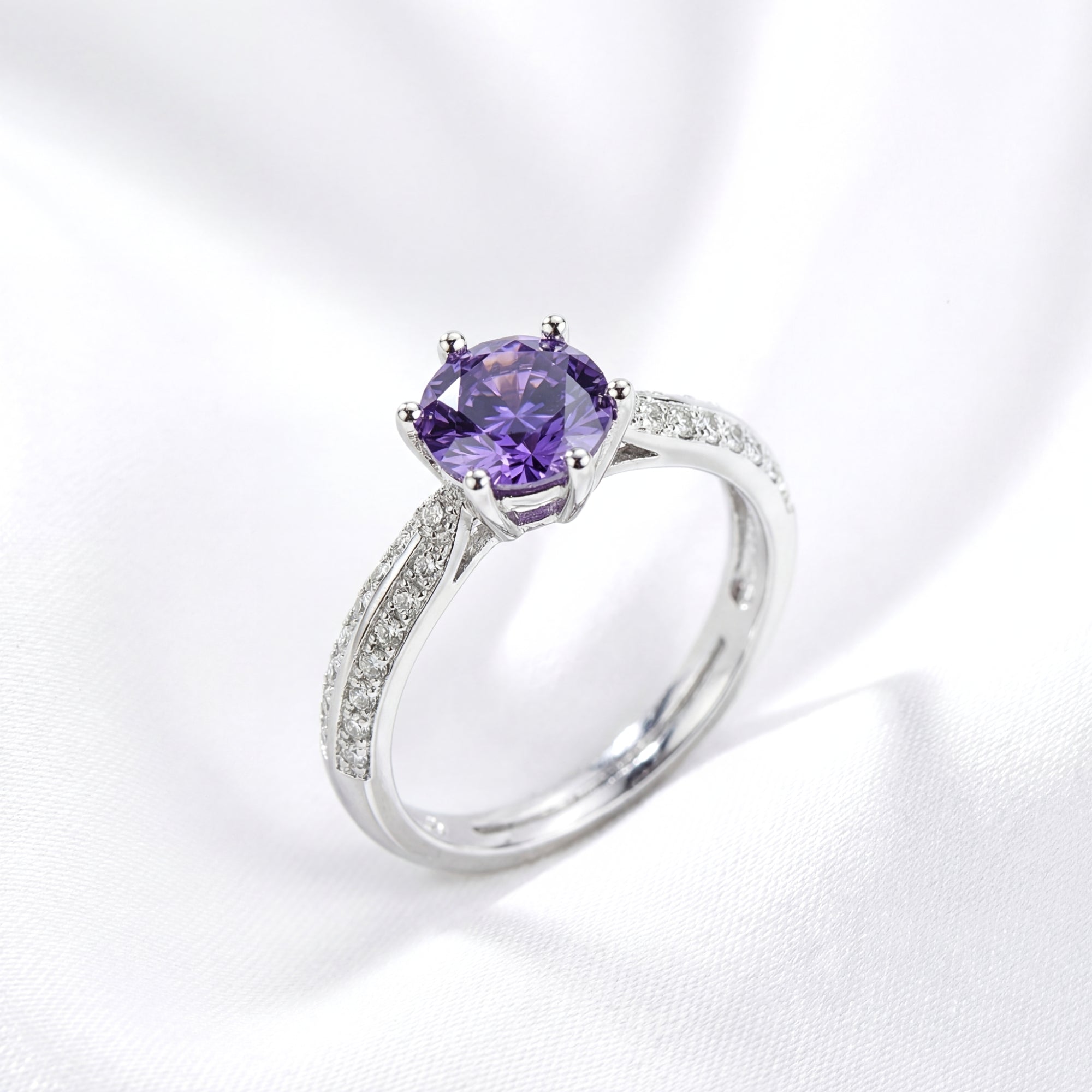 Angled view of a round-cut simulated amethyst solitaire ring in sterling silver on white silk fabric.