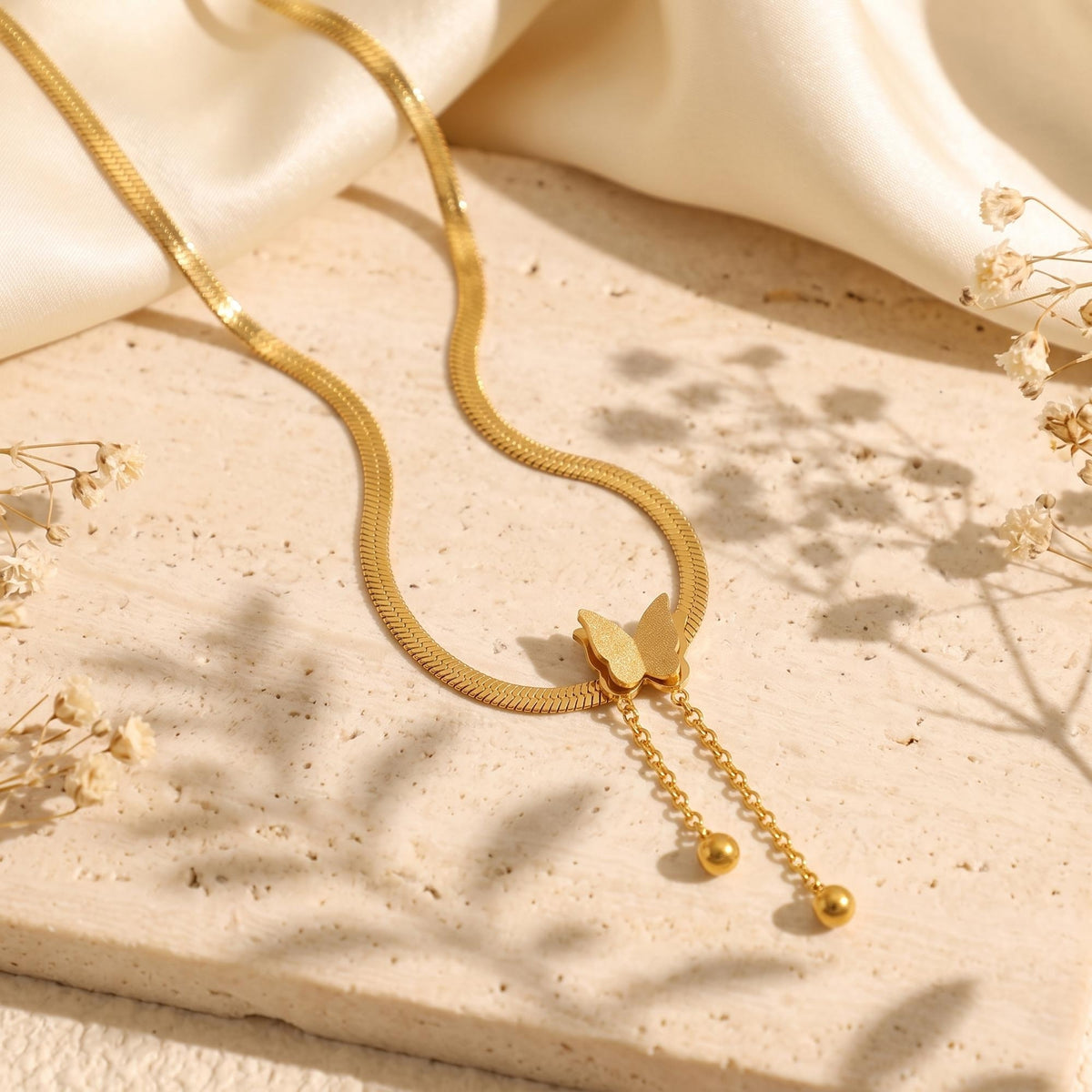 Gold-tone butterfly necklace with a flat chain and two dangling ball-ended chains on a beige stone slab beside cream fabric.