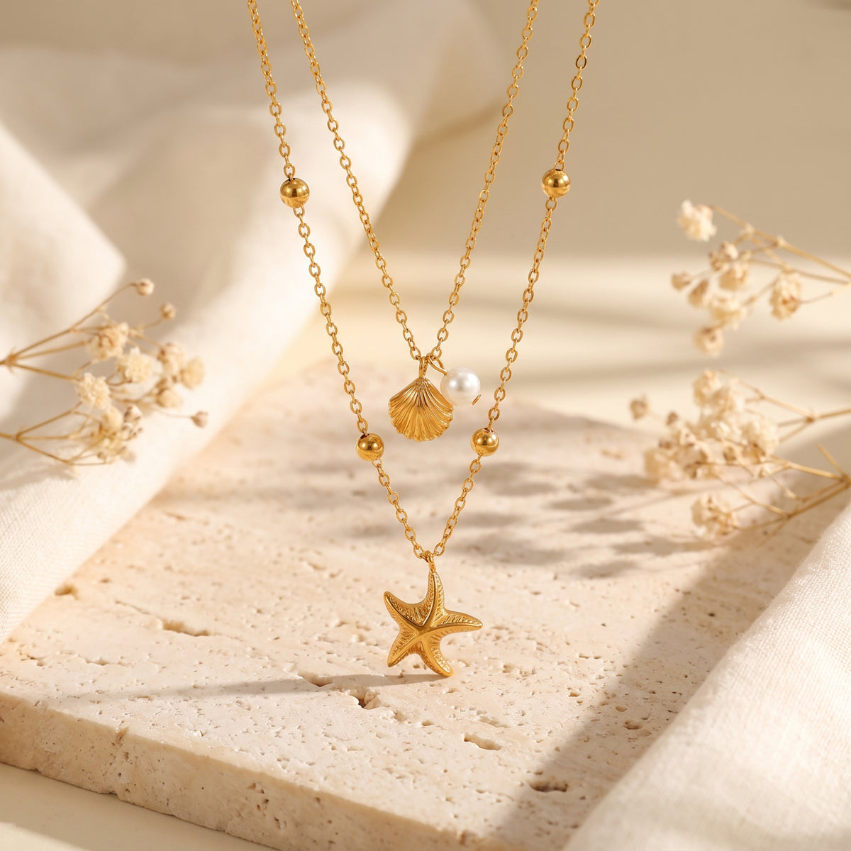 Gold-tone double-layered necklace with shell, simulated pearl, and starfish charms over a textured stone pedestal.