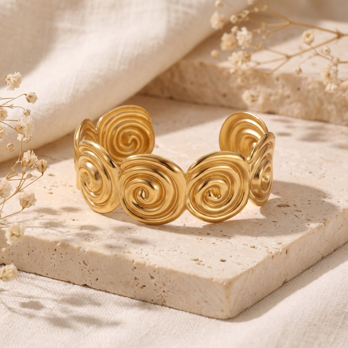Gold-tone open cuff bracelet with sculpted spiral motifs displayed on a porous beige stone block with dried flowers.