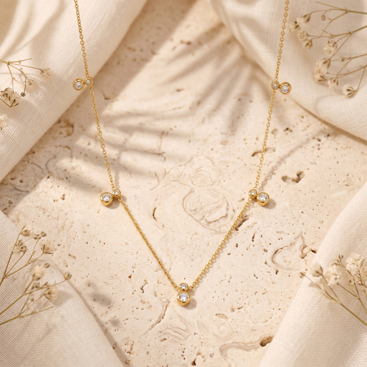 Close-up of the gold-tone station necklace with clear bezel-set stones on a textured beige stone surface.