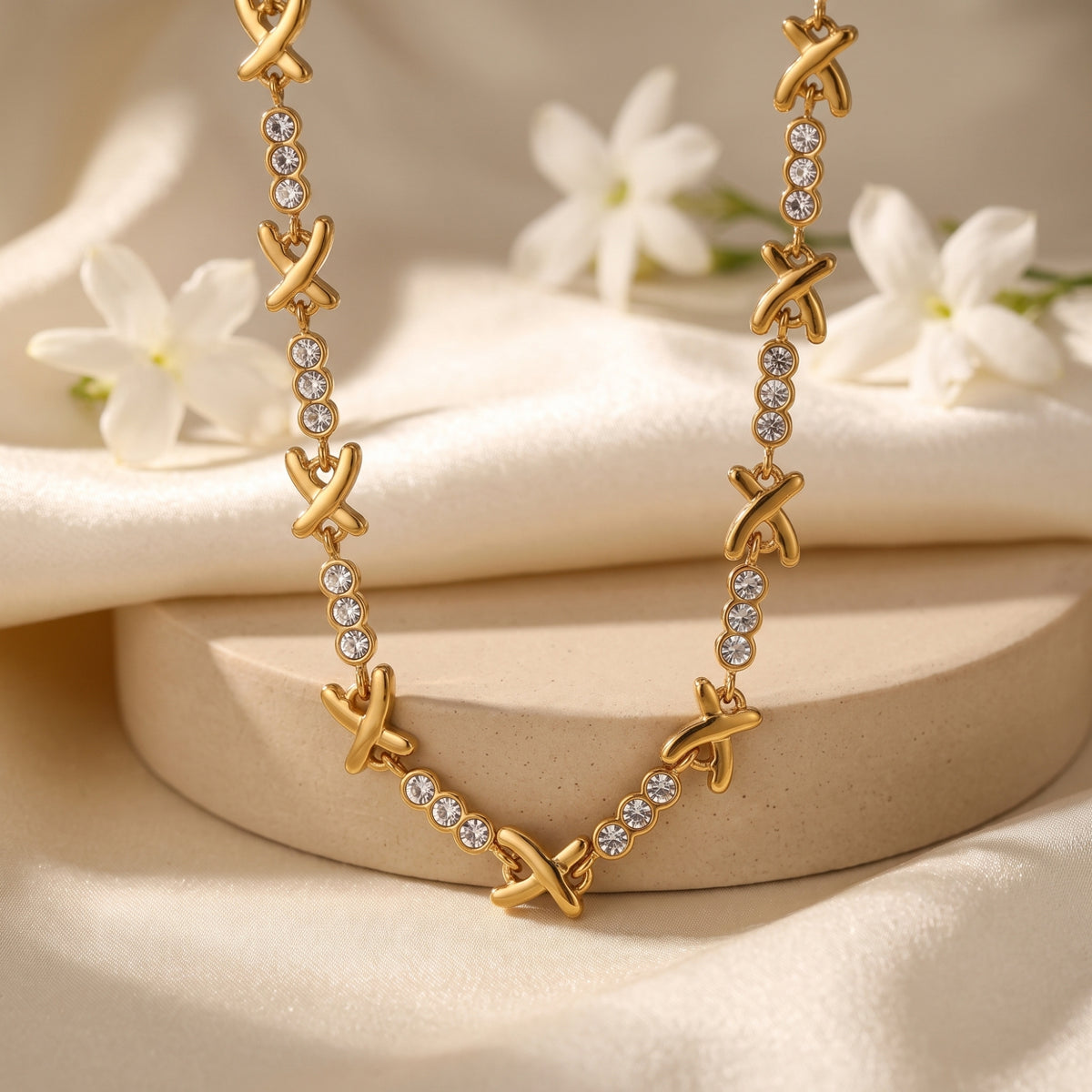 Gold-tone station necklace with X links and round clear stone trios draped over a beige pedestal.