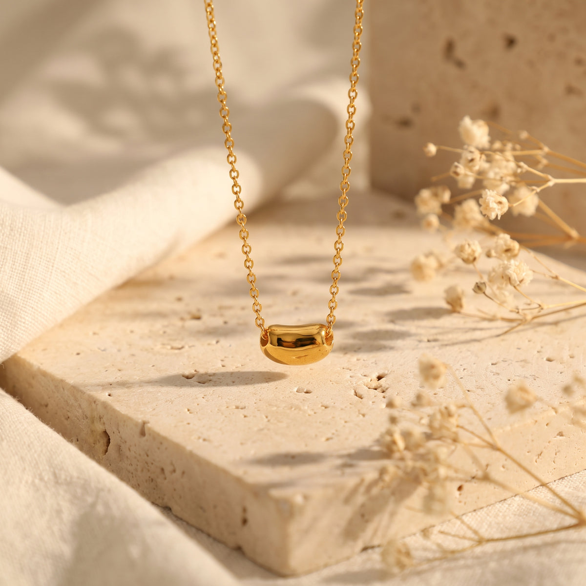 Sculpted gold-toned pod pendant suspended on a fine link chain above a warm stone pedestal.