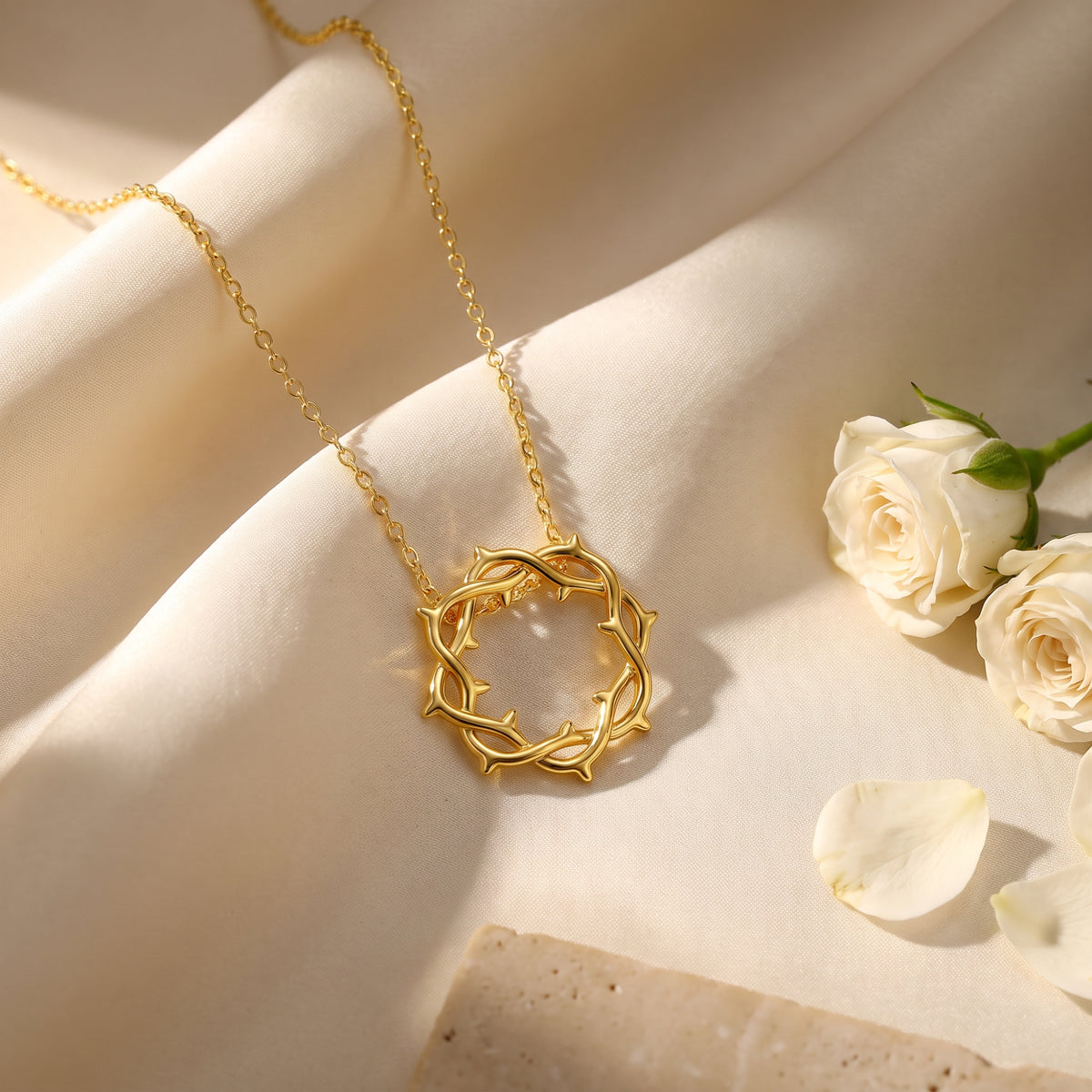 Gold crown of thorns pendant necklace on folded ivory fabric beside white roses and petals.