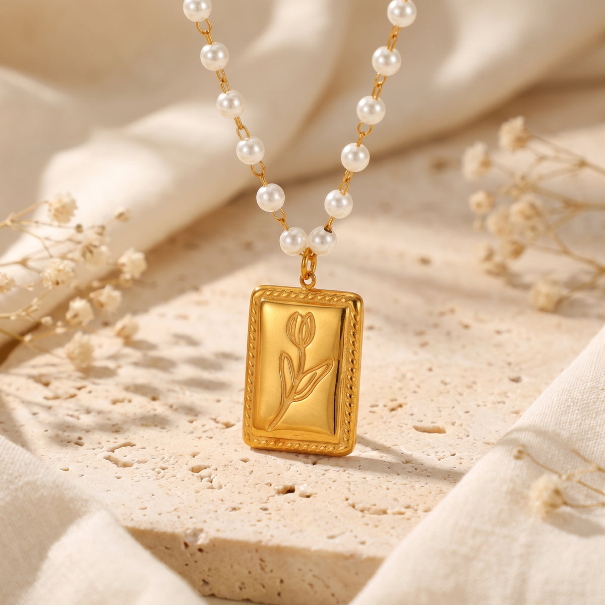 Gold-tone rectangular tulip pendant necklace suspended on a strand of white pearls over beige stone.